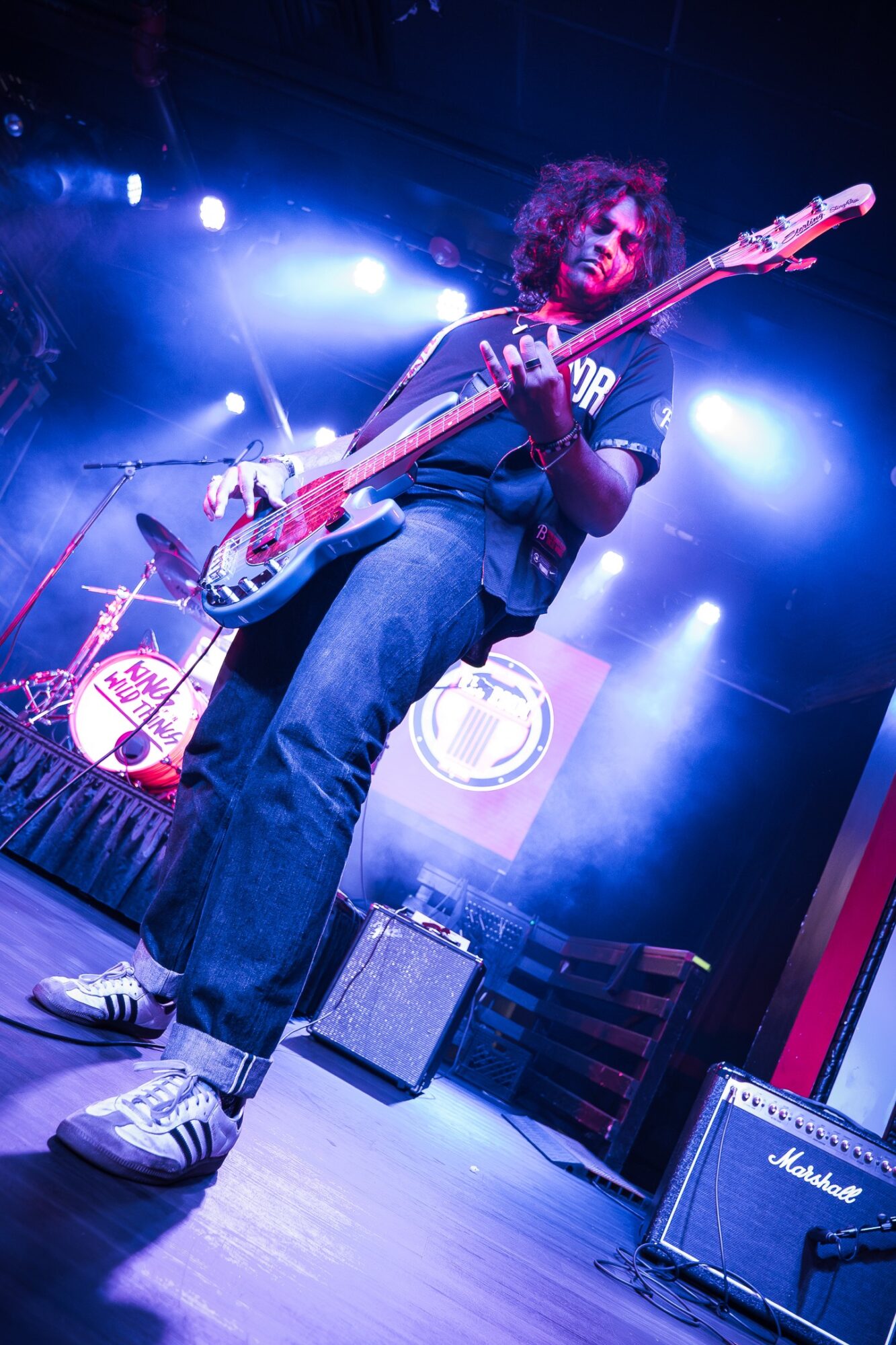 Musician playing bass guitar on stage with blue and purple lighting, wearing a dark shirt and jeans.
