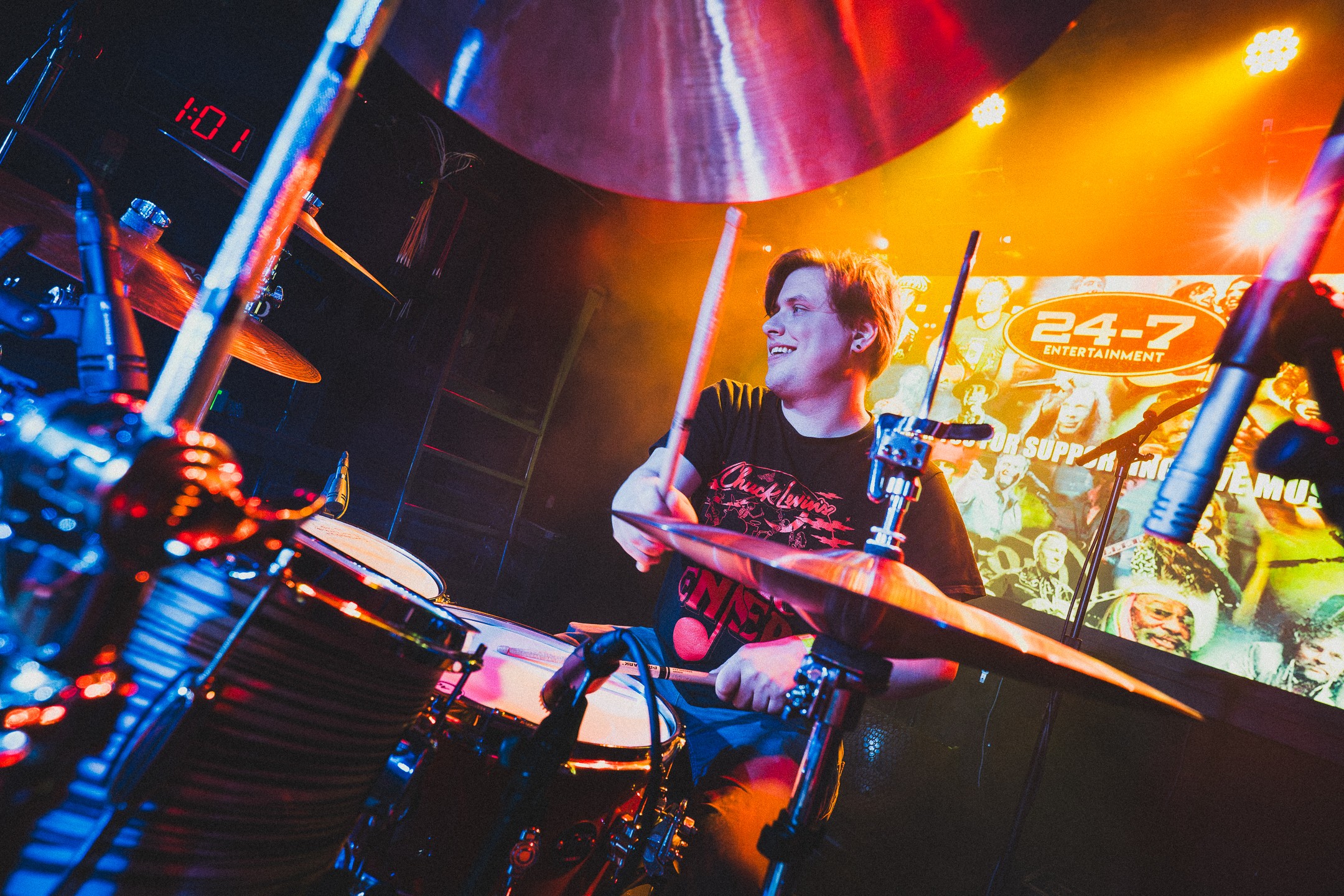 A young woman plays drums on stage with colorful lighting and a screen in the background, smiling and focused.