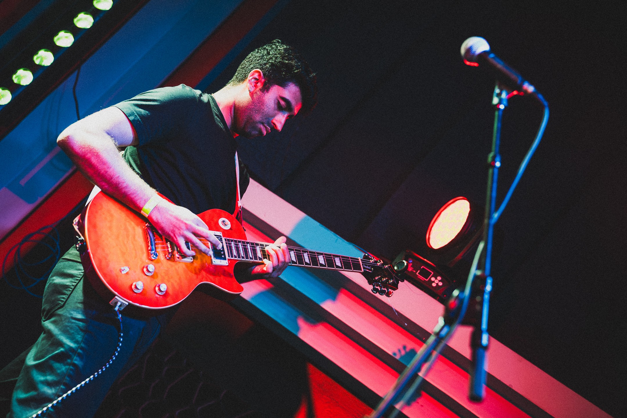 Person playing an orange electric guitar on stage with a microphone stand nearby, dark background, stage lighting.