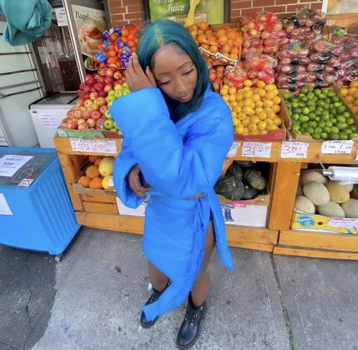 Young woman with blue hair wearing a bright blue coat stands in front of a fruit stand, touching her head.