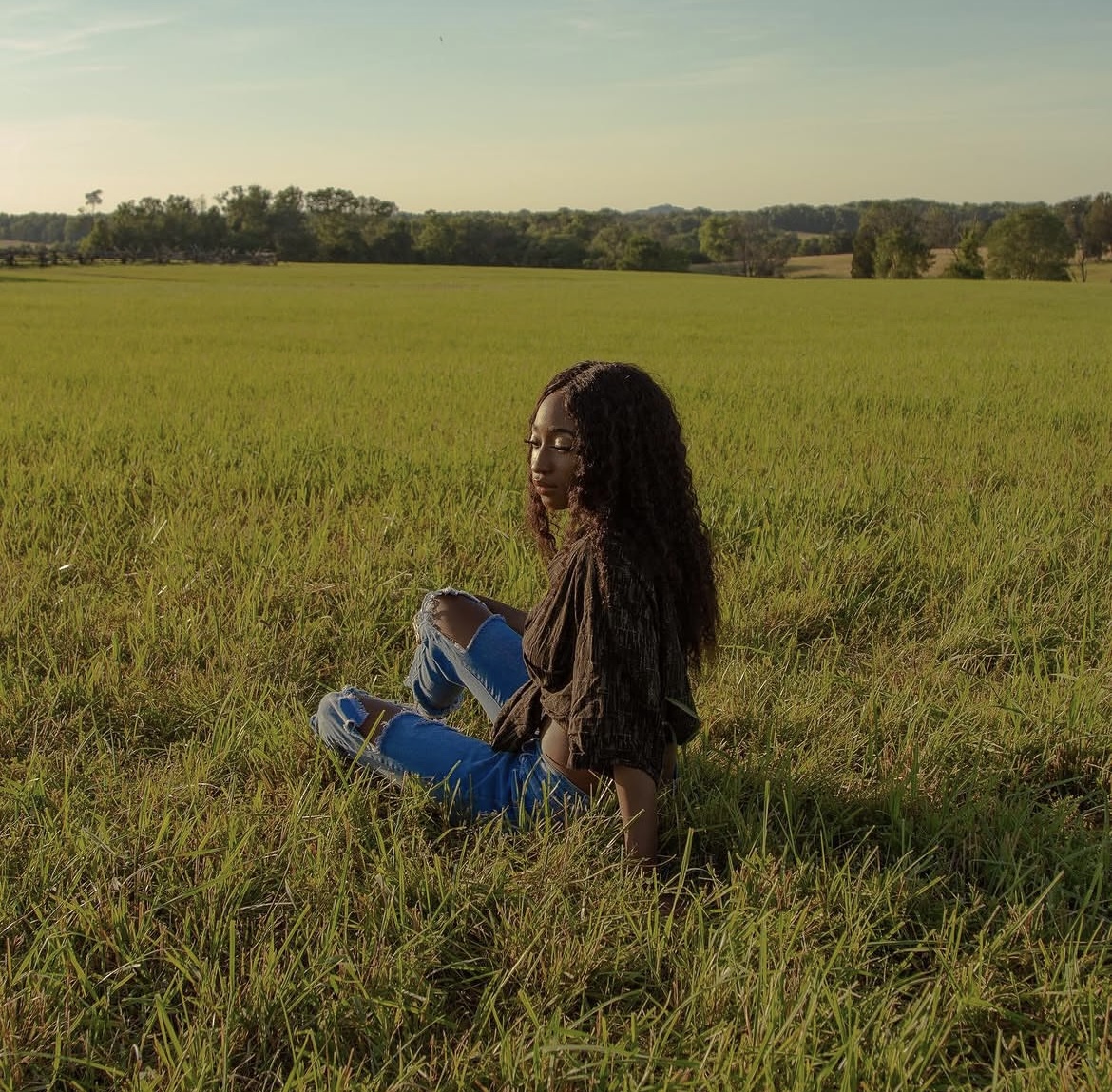 Person sitting on grass in a field, facing right, with trees and sky in the background.