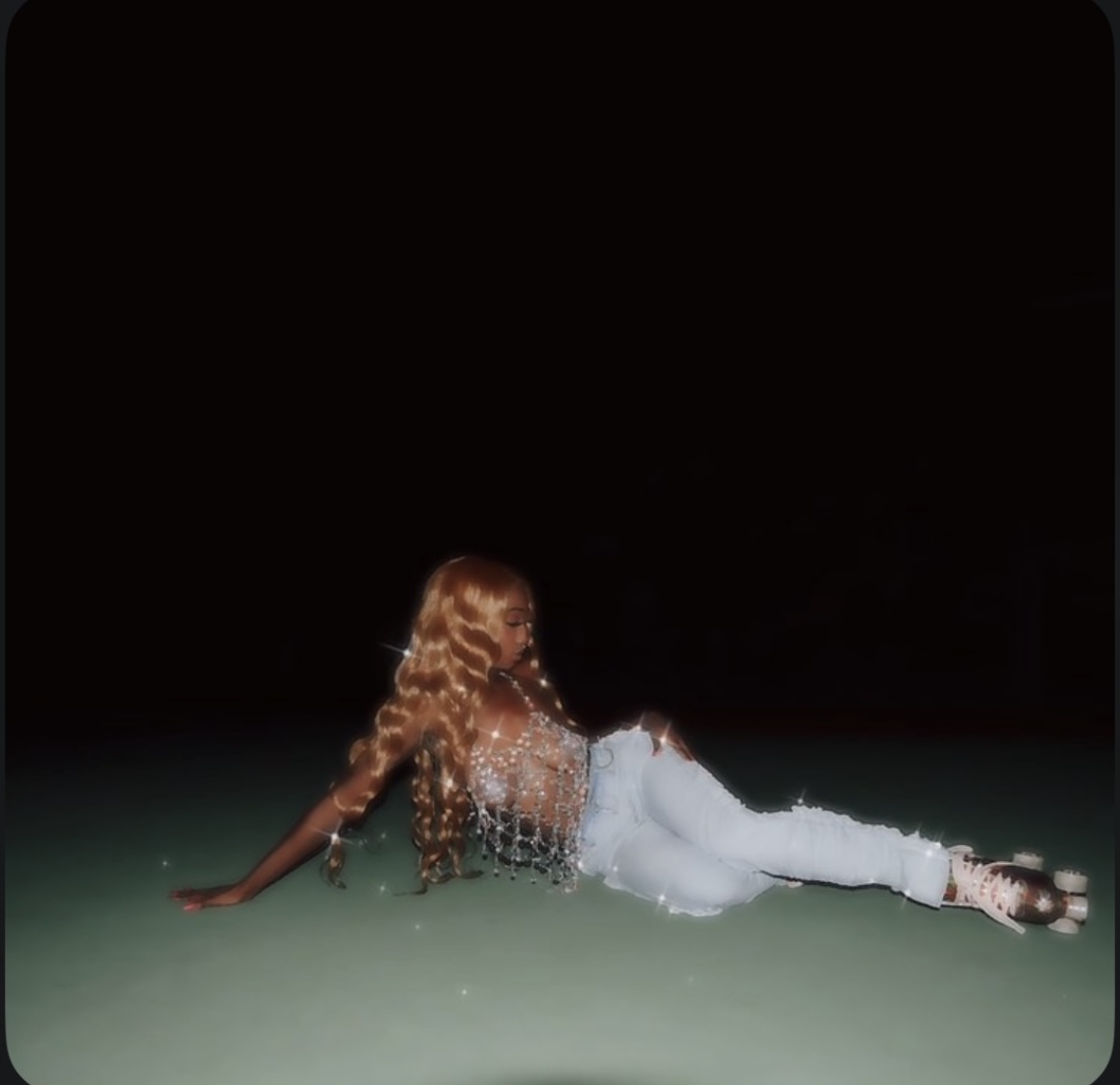Person lying on the ground with long hair, wearing a sparkly top and white pants, against a dark background.