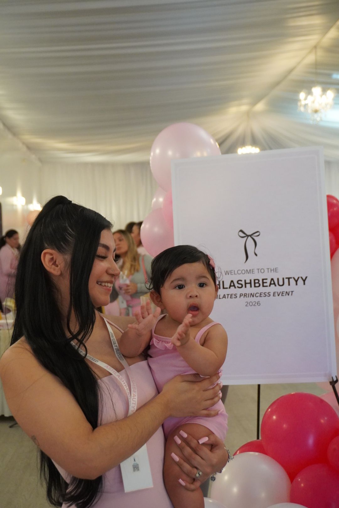 Woman holding a baby girl at an event with balloons and a white sign, smiling and looking at the camera.