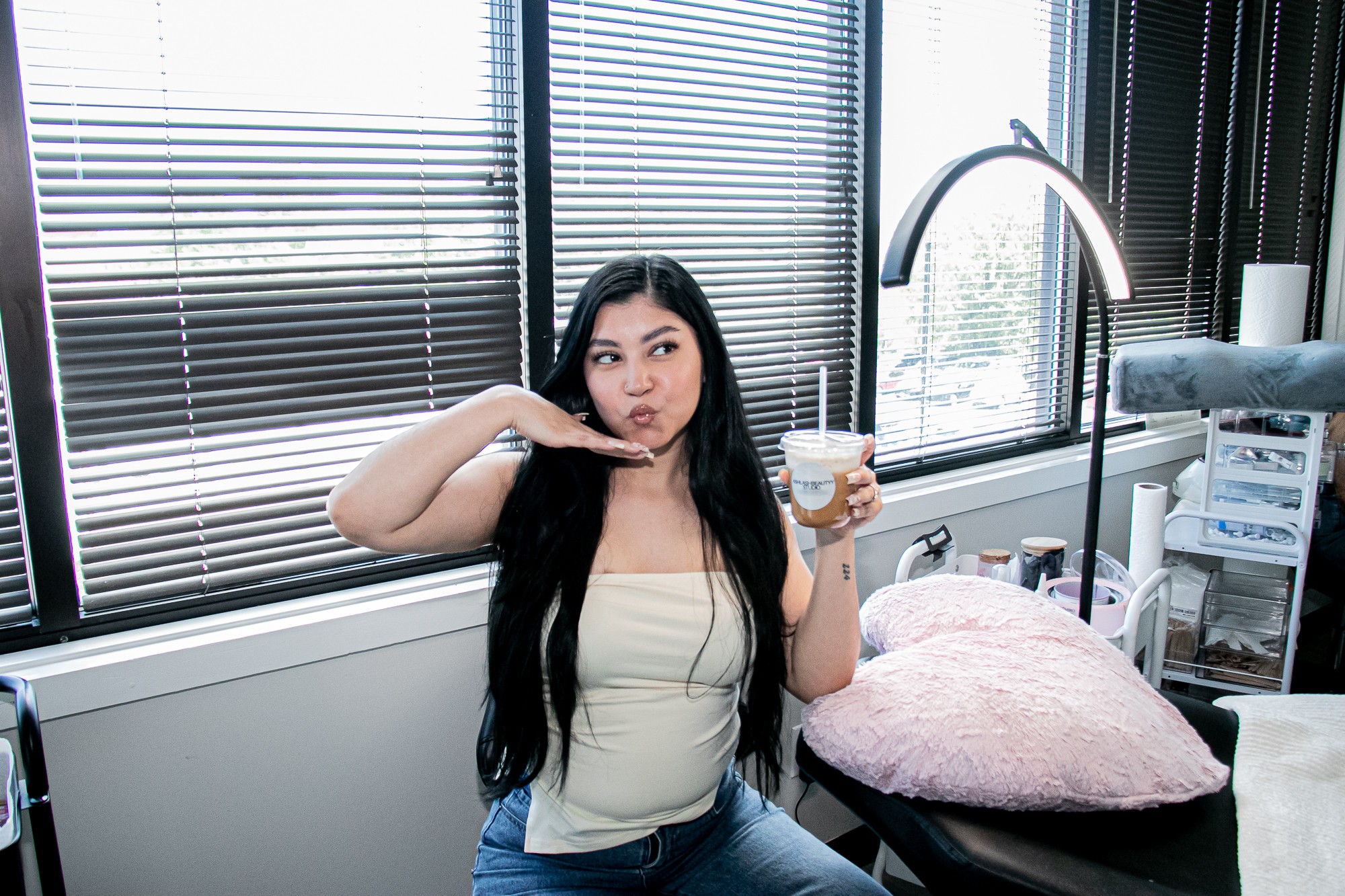 Young woman with long dark hair holding a drink, sitting in a room with large windows and blinds, pink pillow nearby.