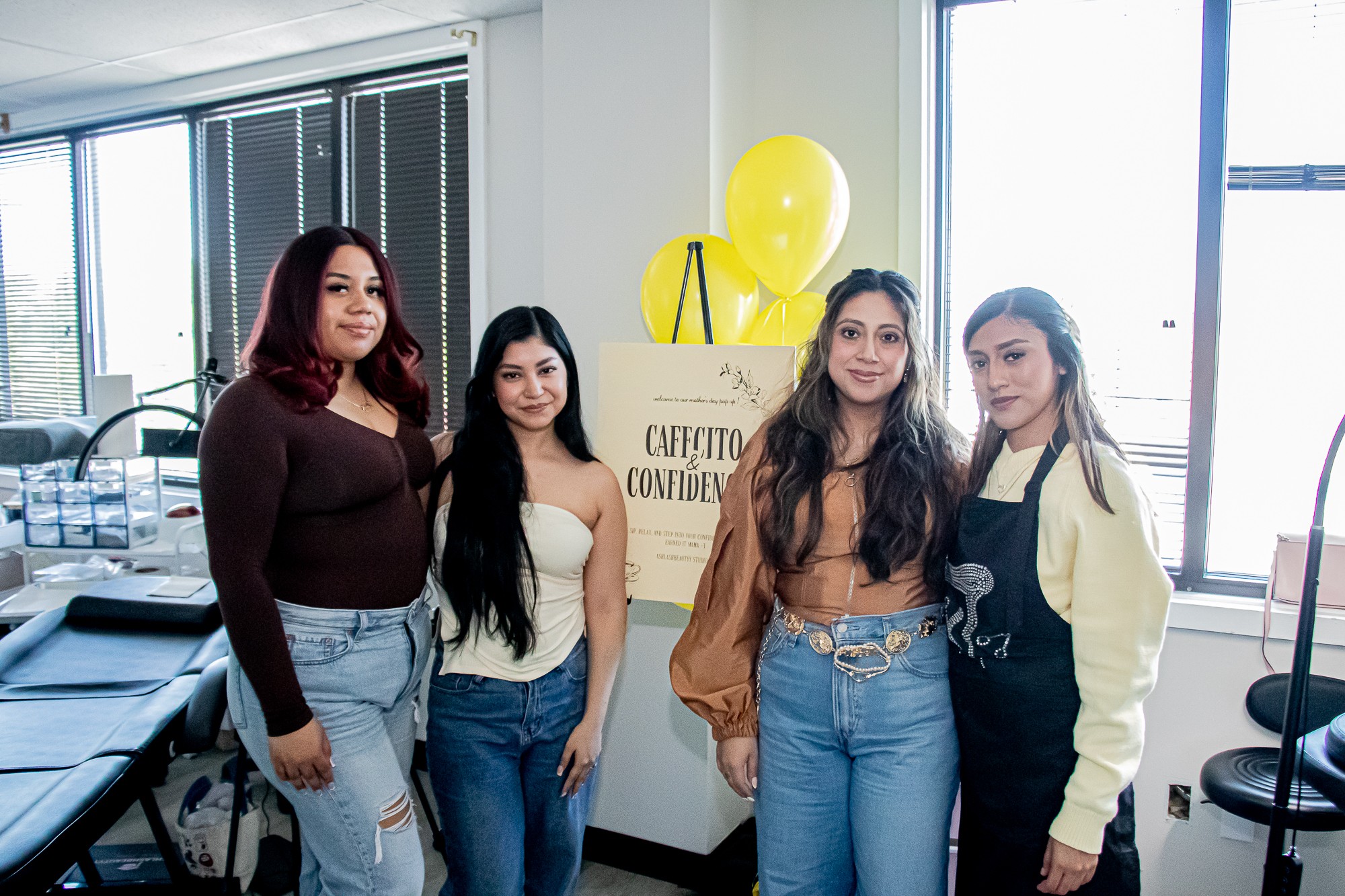 Four women standing together indoors near a window, smiling, with a yellow balloon and a sign in the background.