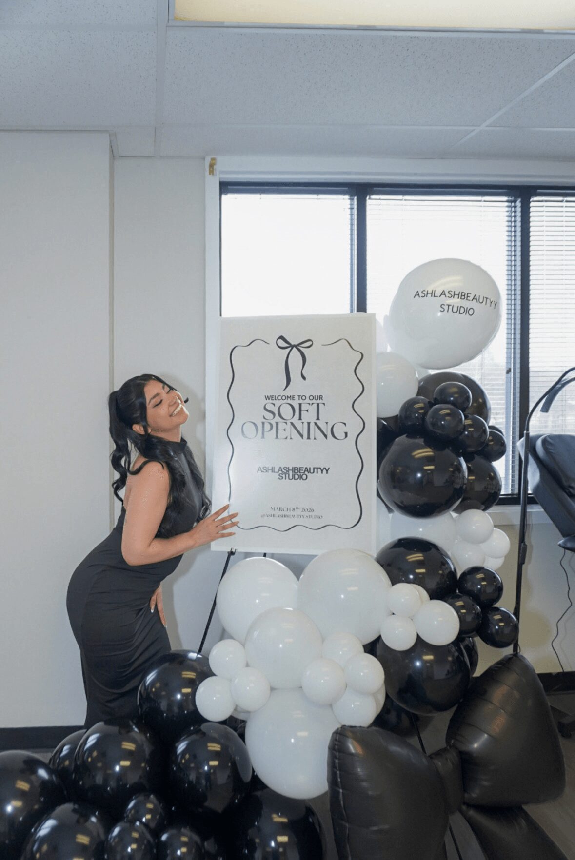 Woman in black dress standing next to a sign for a soft opening, surrounded by black and white balloons, in an office space.
