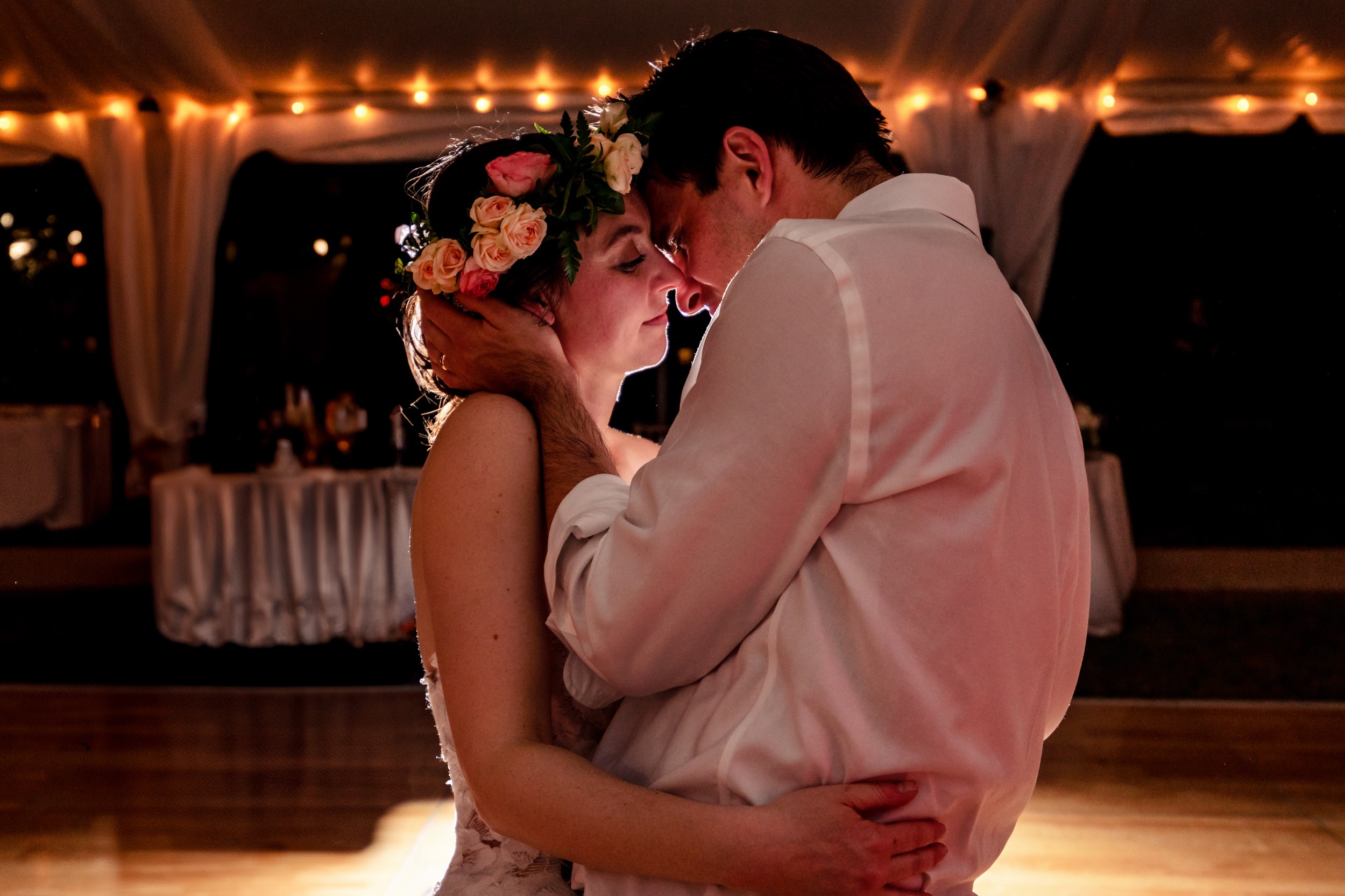 A couple dances closely at night with warm string lights overhead, a decorated table in the background, and a romantic atmosphere.