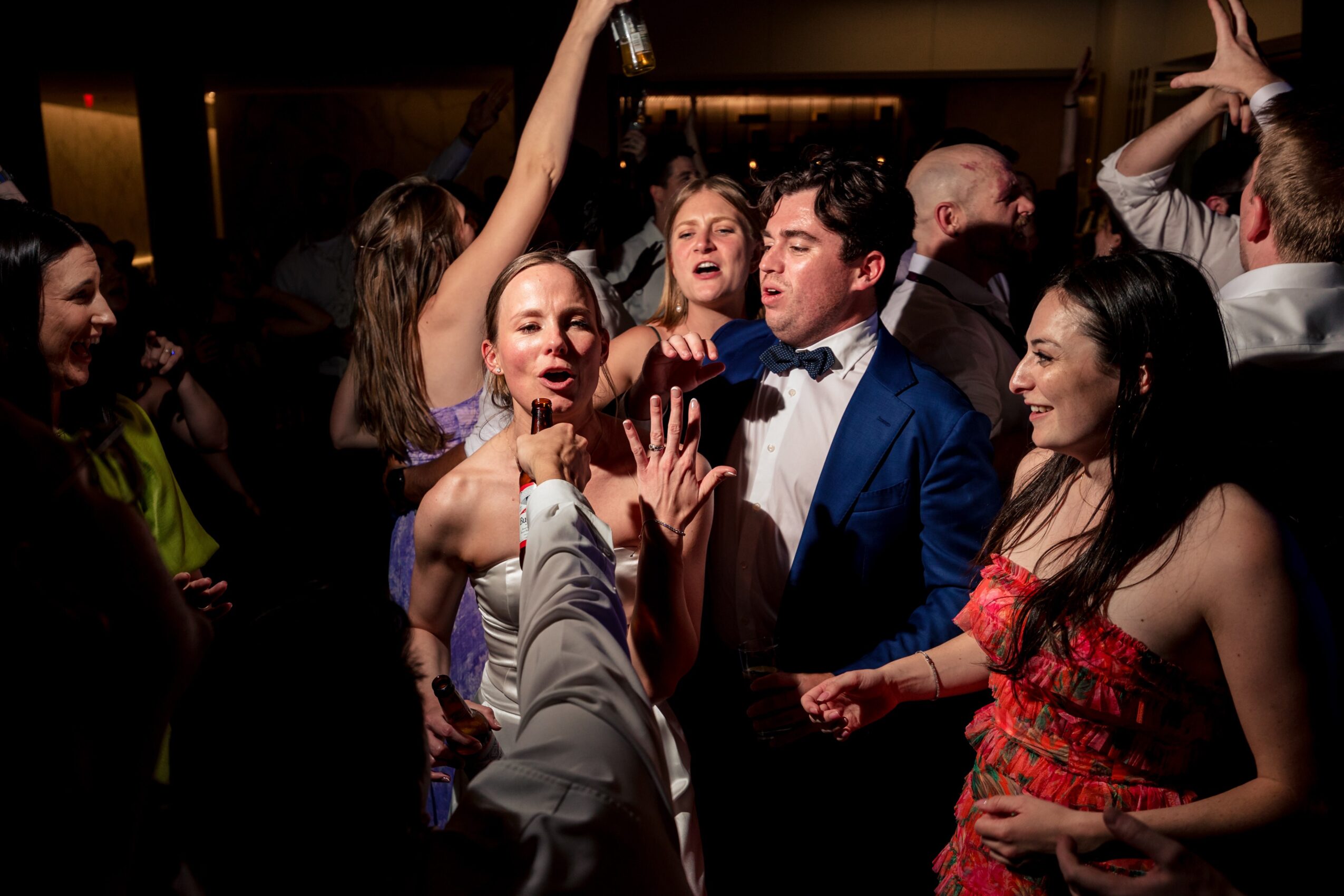 Group of people dancing and socializing at a party, some smiling and raising hands, in a dimly lit room.