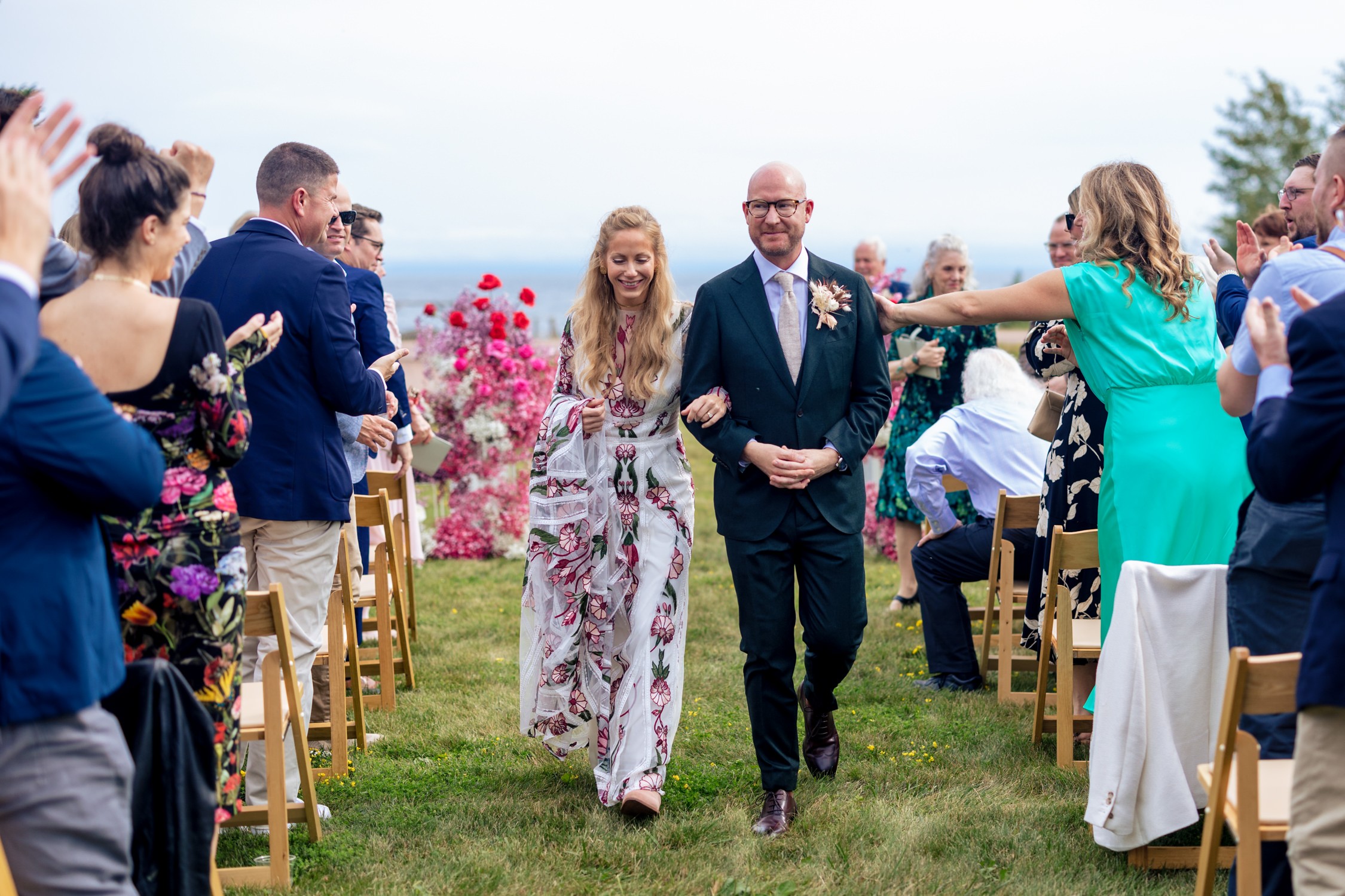 People at an outdoor wedding ceremony, with a woman in floral dress walking with a man in a suit, guests seated on sides.