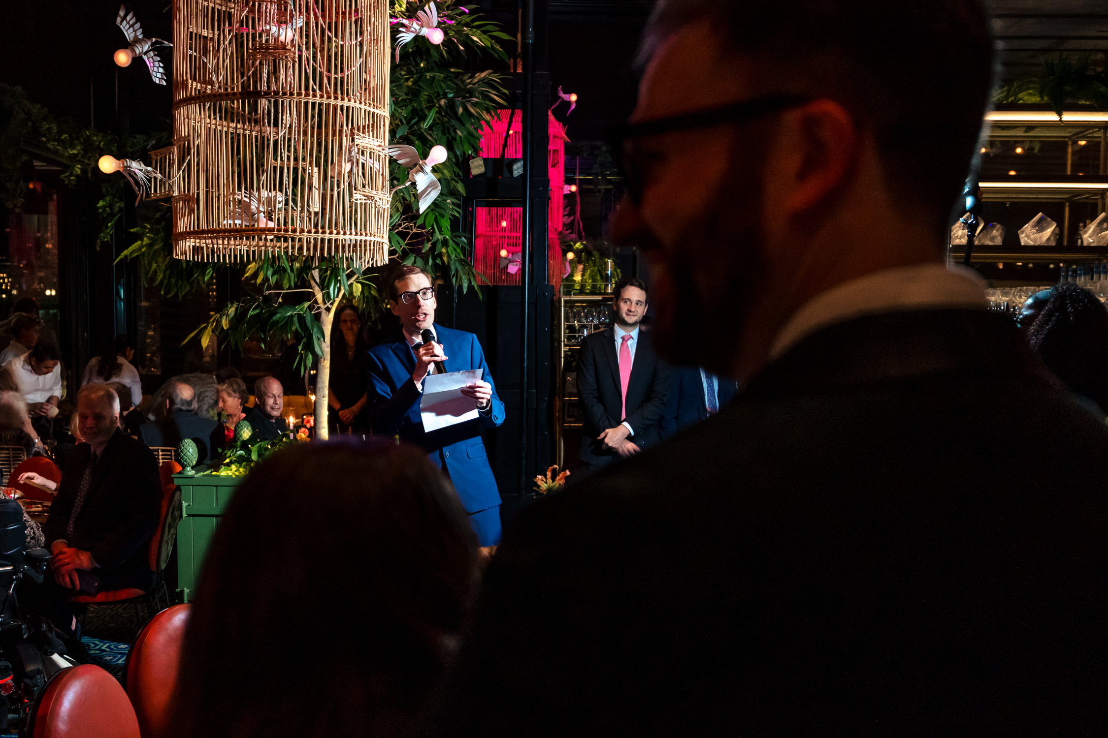 People gathered at an indoor event with decorative lighting and plants, some speaking or listening, in a lively setting.