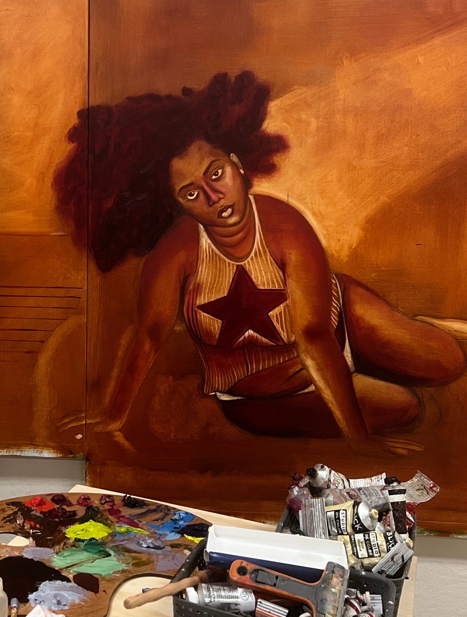 Painting of a woman with large curly hair, sitting against a warm-toned background, wearing a striped top with a star design.