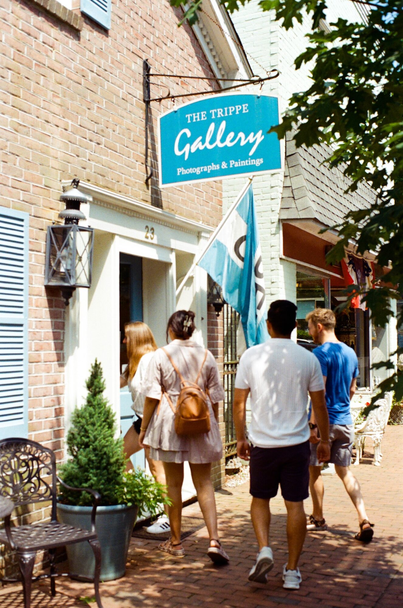 Group of people walking past a gallery sign on a brick building sidewalk, with trees and outdoor seating nearby.