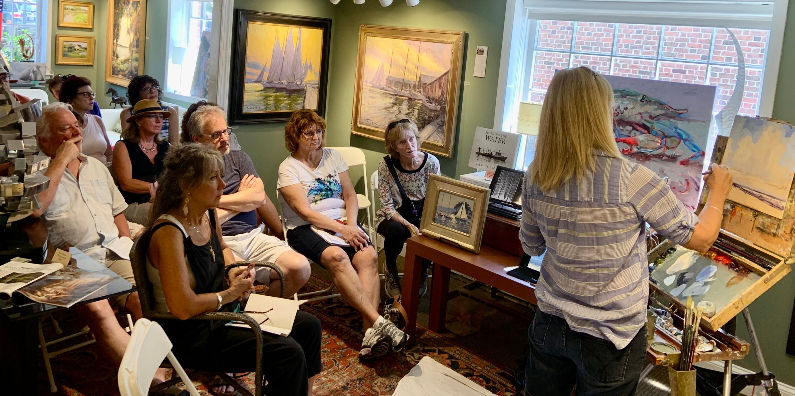 Group of people seated in an art gallery, listening to a woman presenting paintings on an easel.