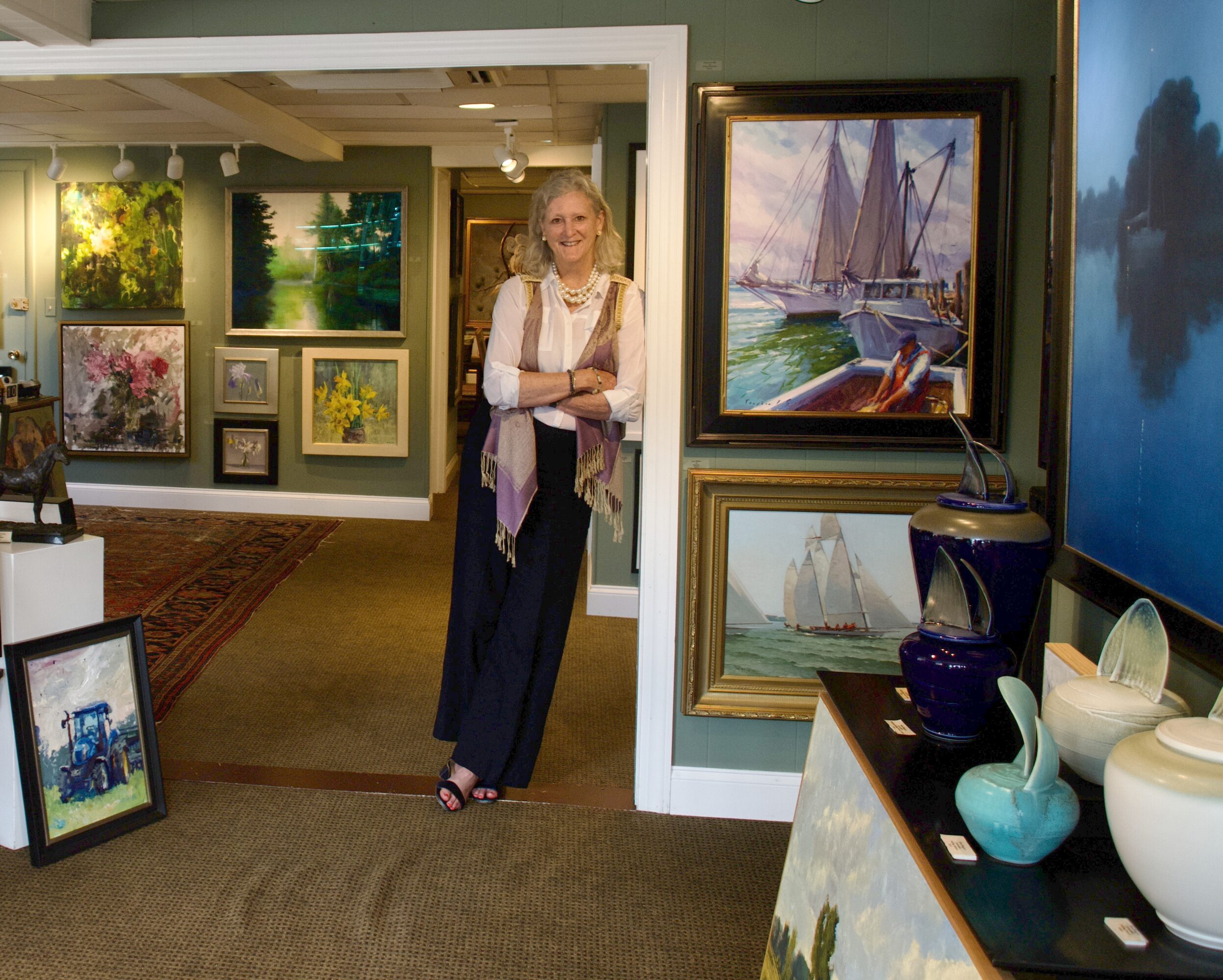 Woman standing in an art gallery with paintings on walls and display shelves, smiling, with a carpeted floor and green walls.