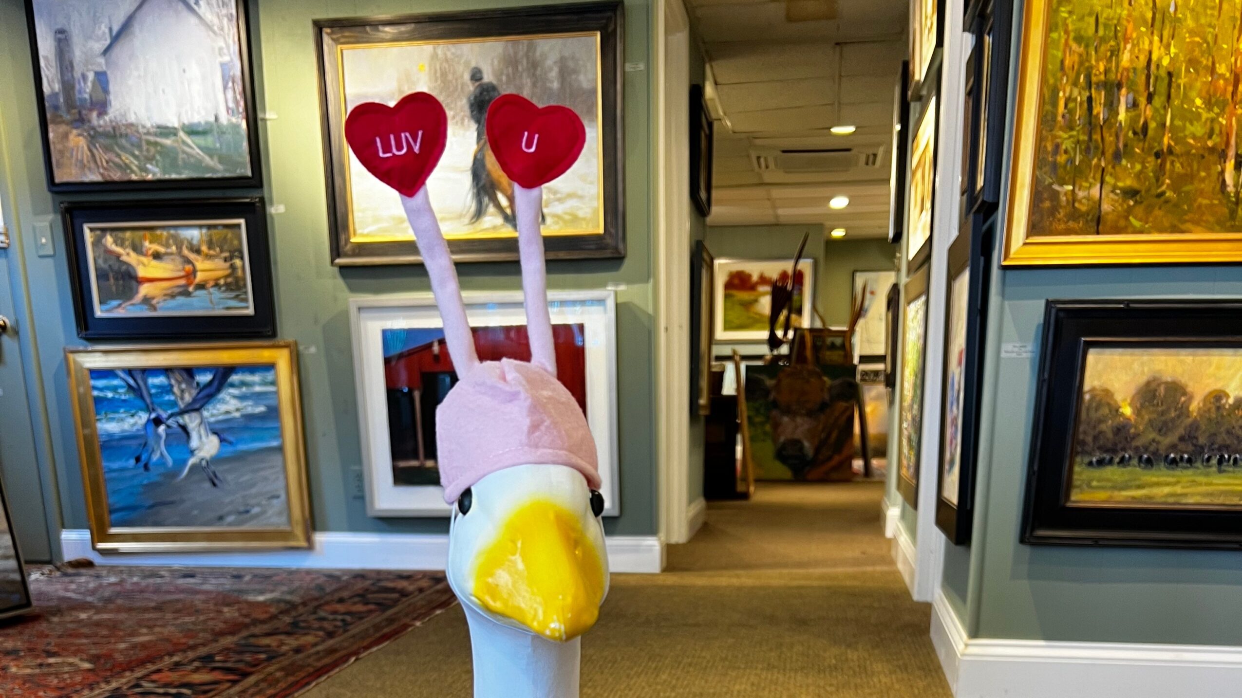 A swan sculpture with pink antennae and red hearts on top, in an art gallery with framed paintings on walls.