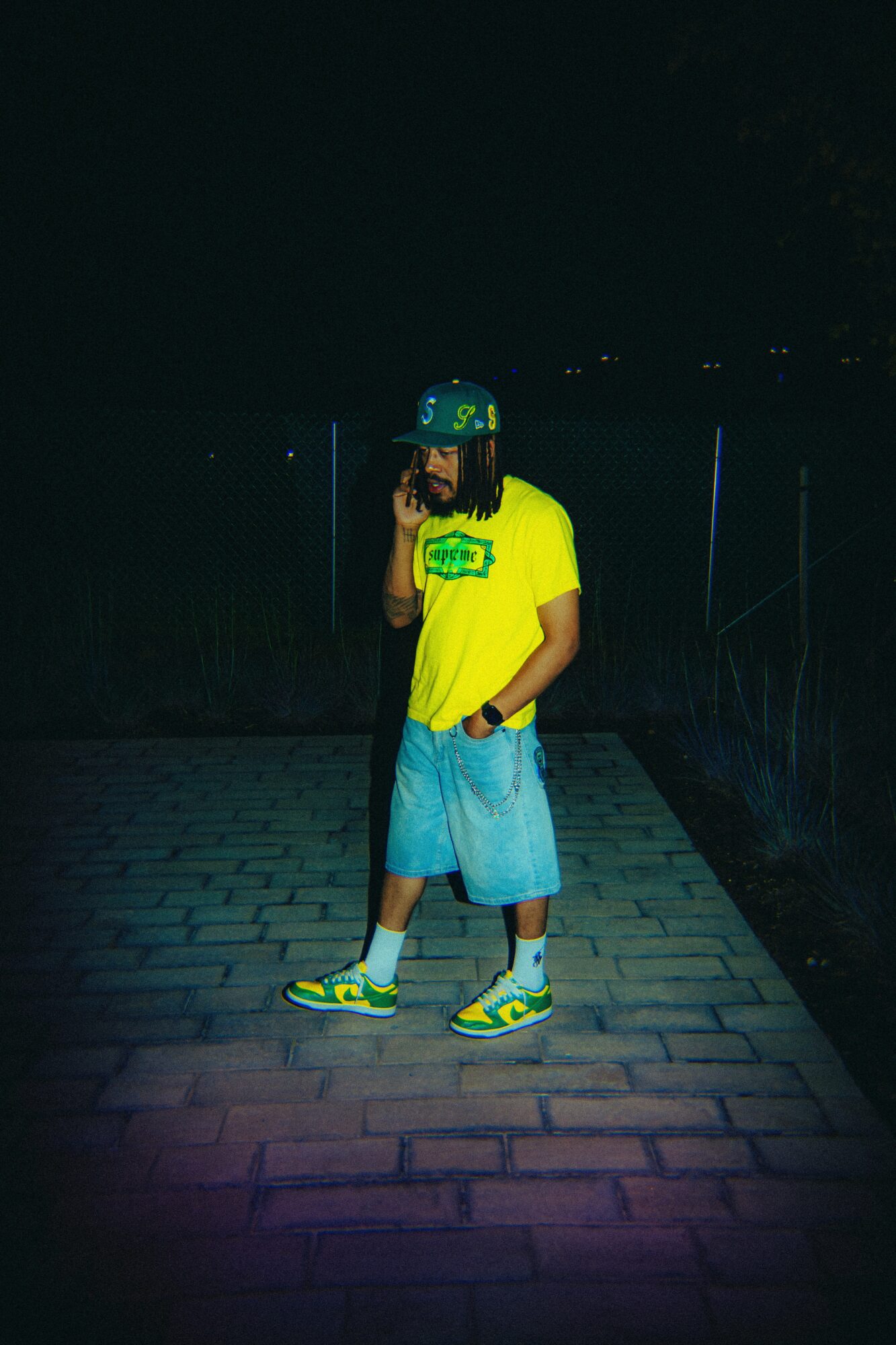 Person standing outdoors at night, wearing a bright yellow shirt, blue shorts, and colorful sneakers, holding a phone to their ear.