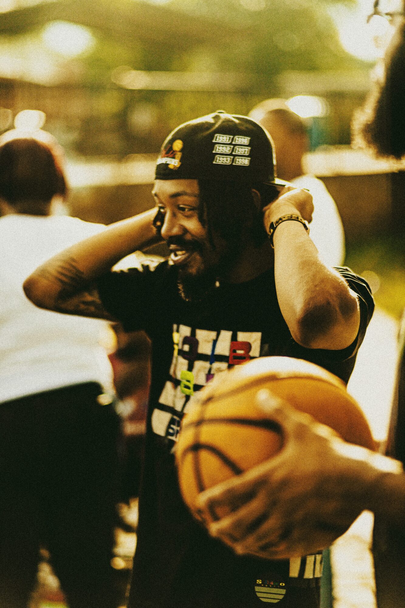 Person smiling, wearing a cap backwards, holding a basketball, outdoors with blurred background of people and trees.