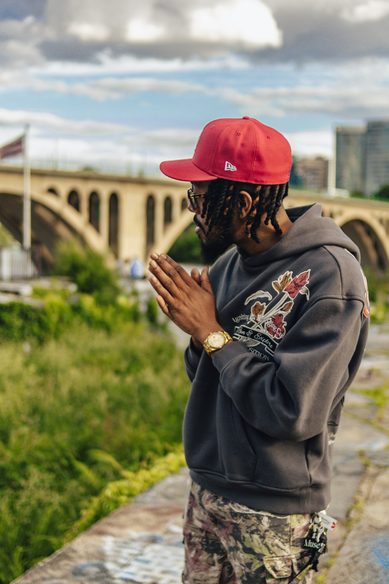 Person wearing a red cap and gray hoodie with hands together outdoors, with a bridge and cityscape in background.