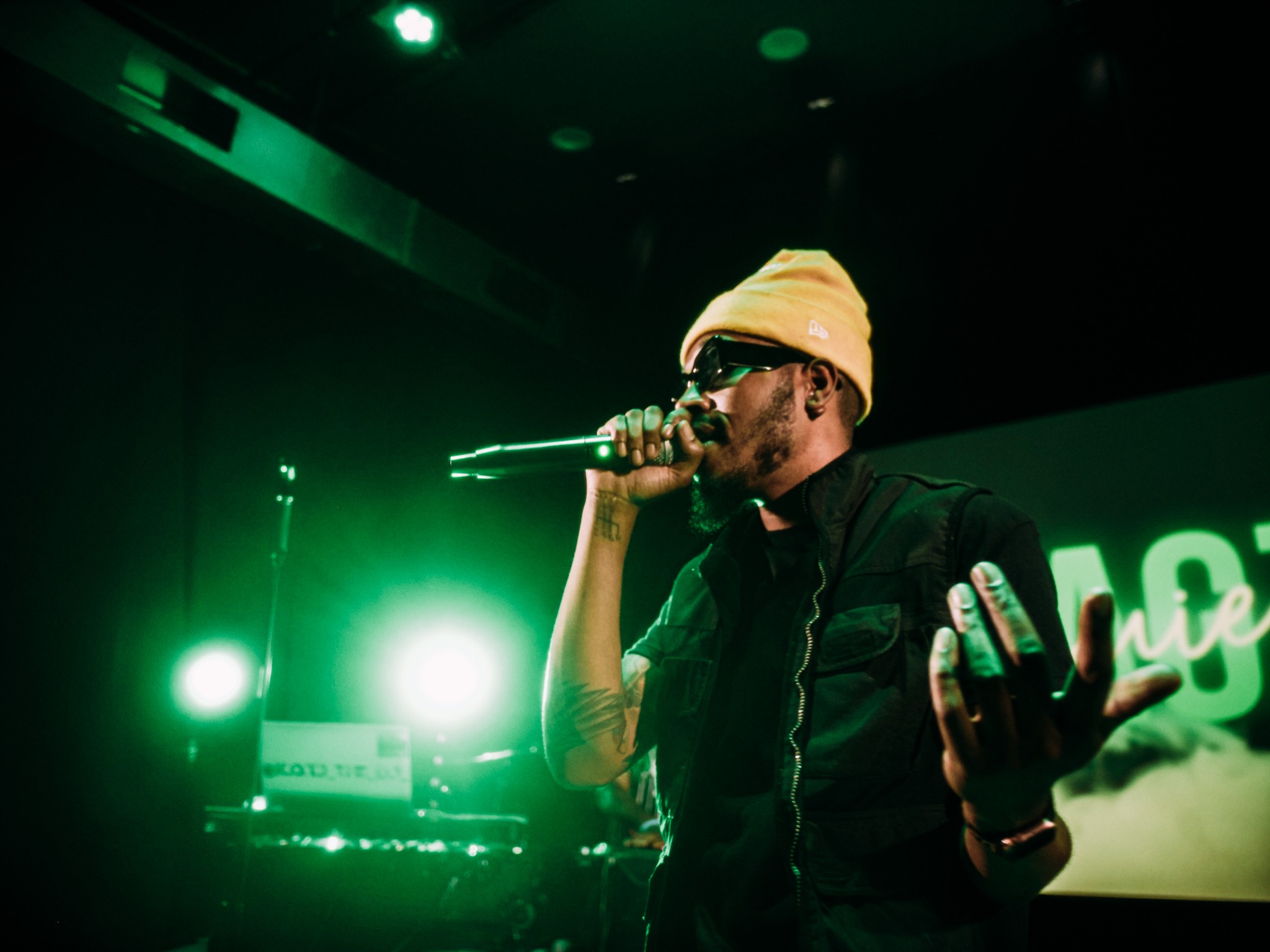 Performer wearing a yellow beanie and sunglasses singing into a microphone on stage with green lighting, holding a phone.