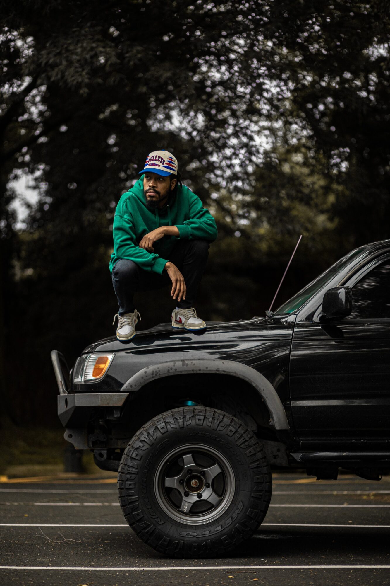 Person crouching on the hood of a black vehicle outdoors, surrounded by trees, wearing a green hoodie and a cap.