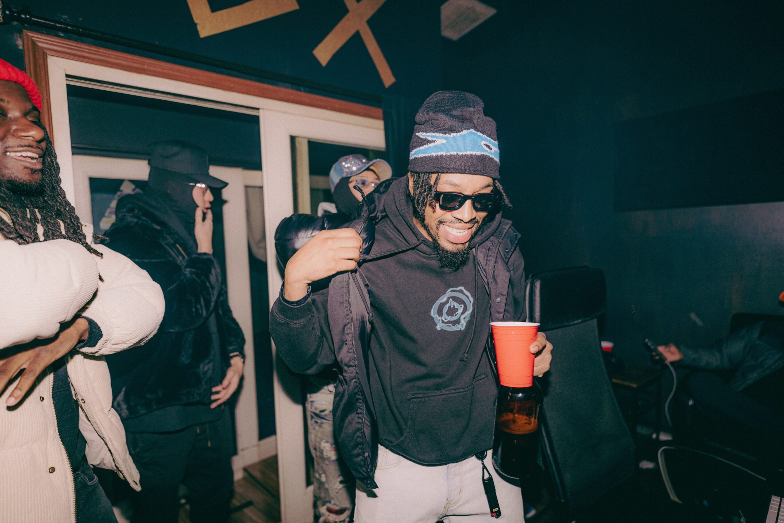Group of people at a party, smiling and holding drinks, with a person in sunglasses and a beanie in focus.