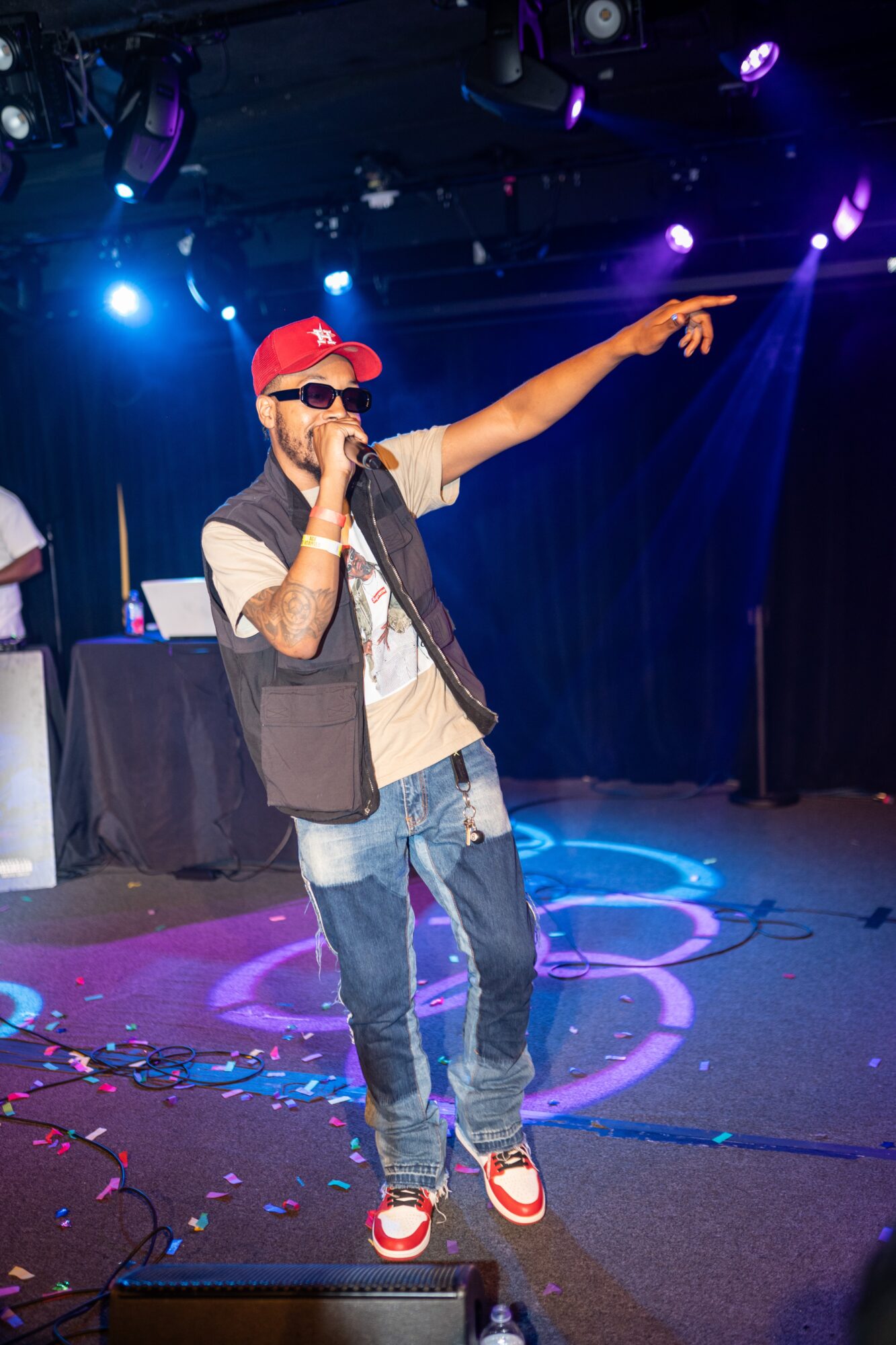 Person performing on stage with microphone, wearing sunglasses, red cap, and casual clothing, pointing to the right, colorful stage lights.