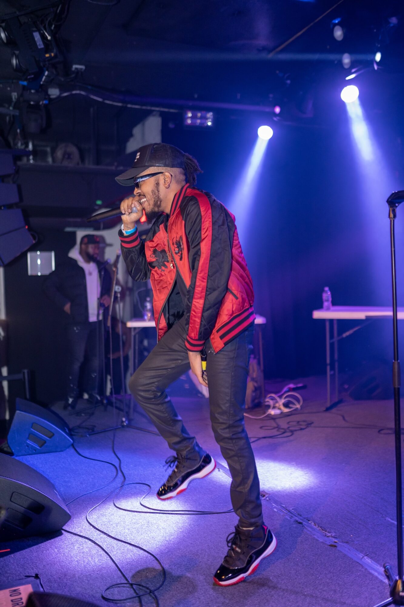 Performer singing on stage with microphone, wearing a red and black jacket, jeans, and a cap, with stage lights shining.