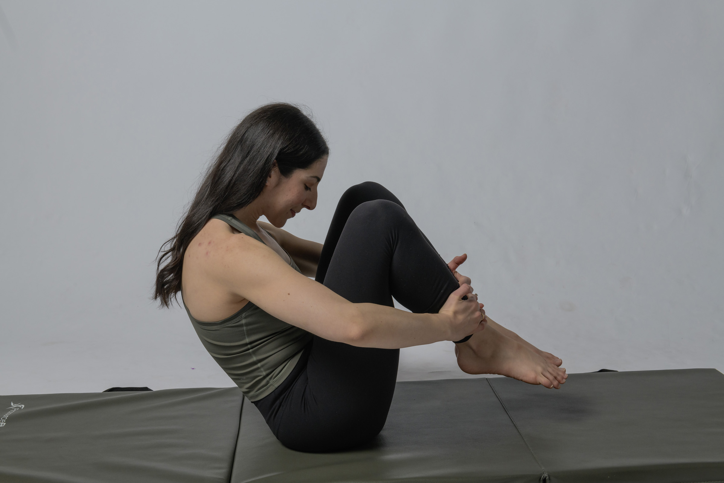 Woman in a yoga pose on a mat, holding her legs close to her chest, smiling, with a plain background.