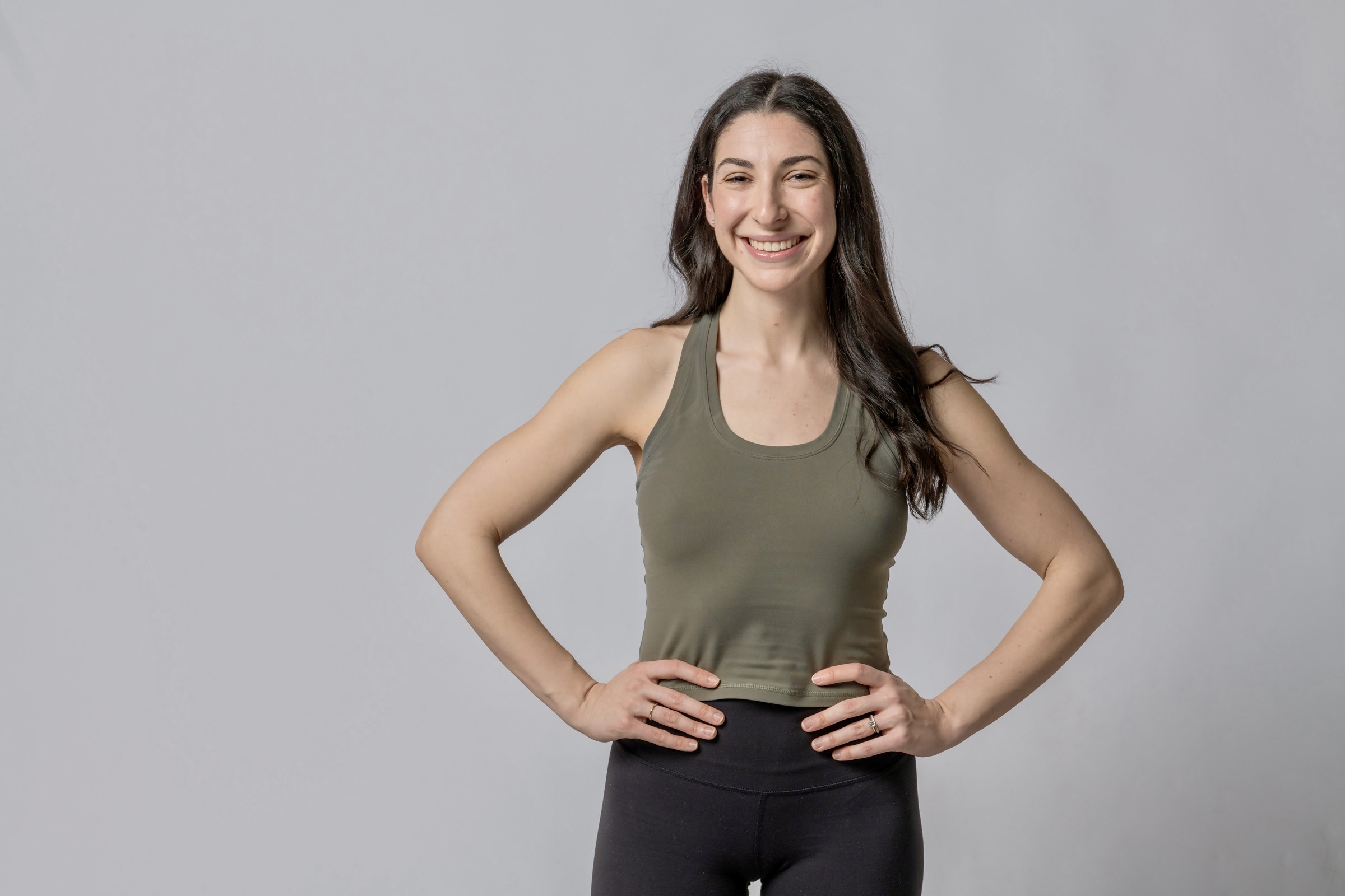 Smiling woman with long dark hair wearing a green tank top and black pants, hands on hips, standing against a gray background.