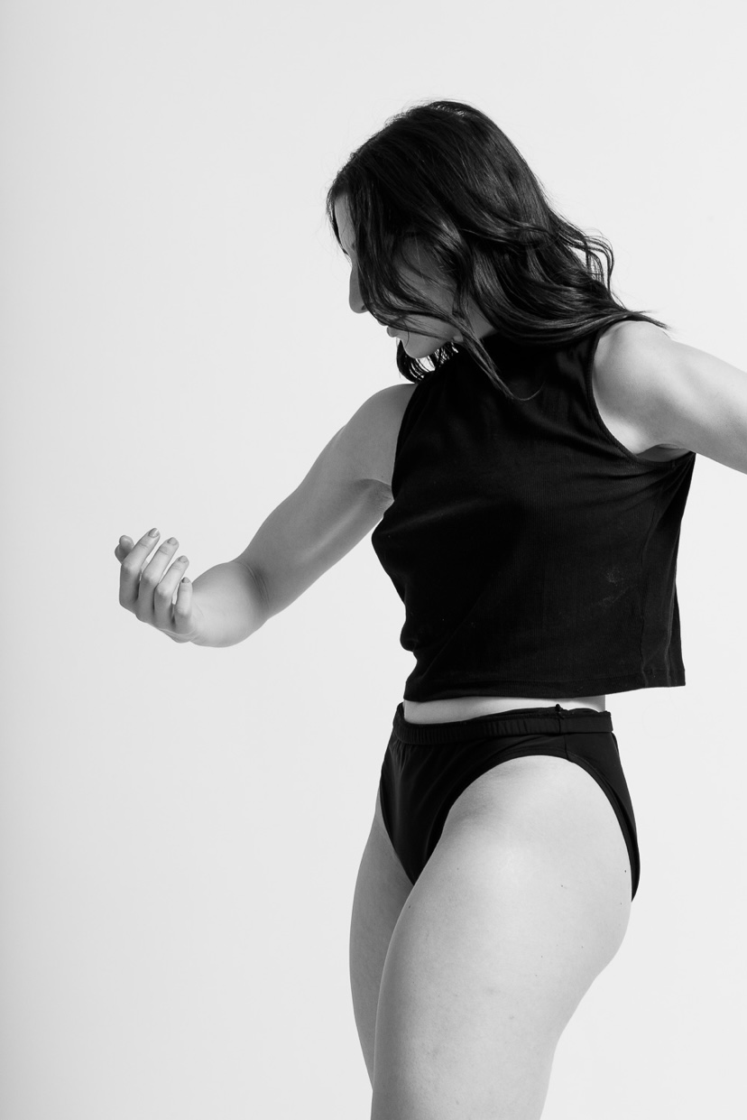 Woman in black sleeveless top and underwear looking down, flexing her arm, against a plain background.