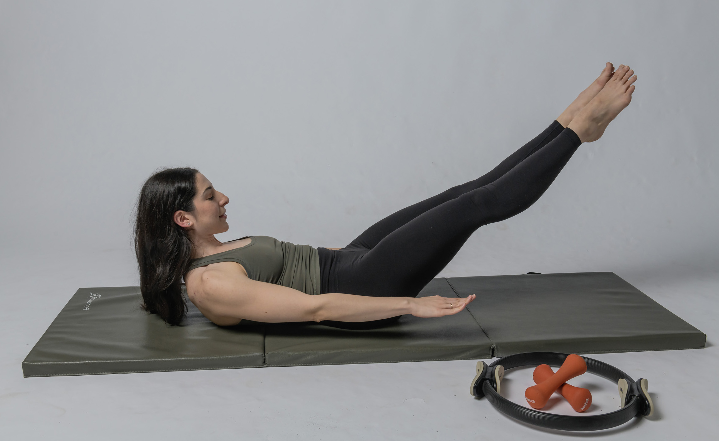 Woman lying on her back on a mat, legs raised and extended, performing a Pilates exercise, with small dumbbells nearby.