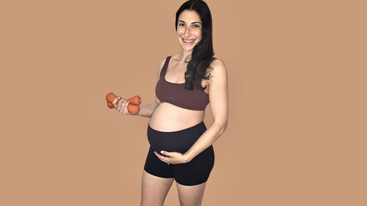Pregnant woman holding small dumbbells and smiling, standing against a plain background.