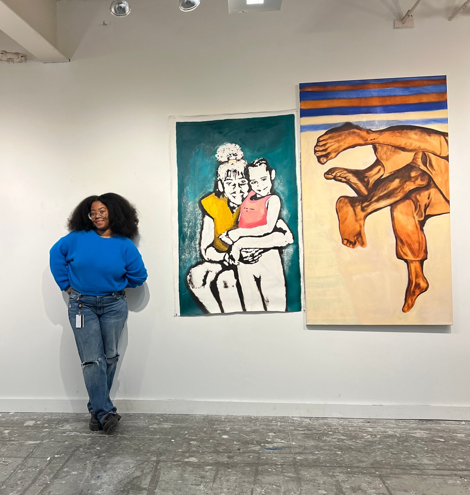Woman with curly hair in a blue sweater and jeans standing next to two paintings on a white wall.