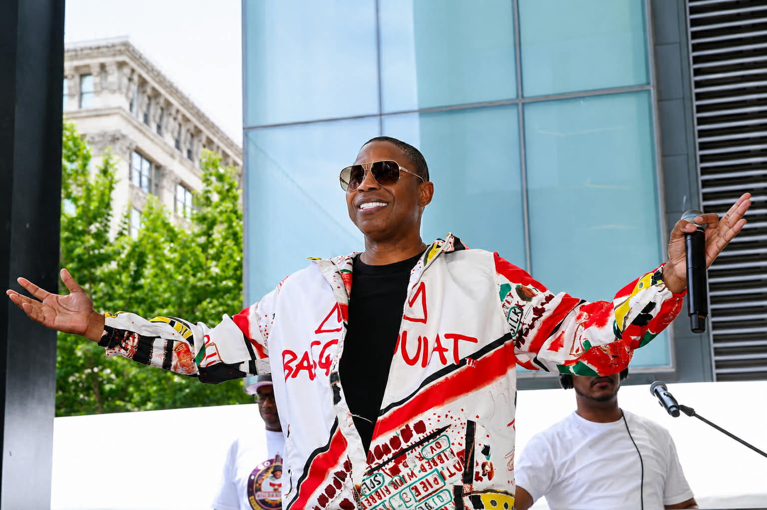 Person wearing sunglasses and a colorful jacket with arms outstretched outdoors, modern building in background.