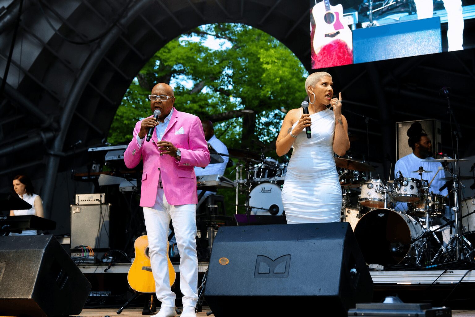 Two men with microphones on stage at an outdoor event, one in a pink blazer and white pants, the other in a white dress, with band equipment and trees in background.