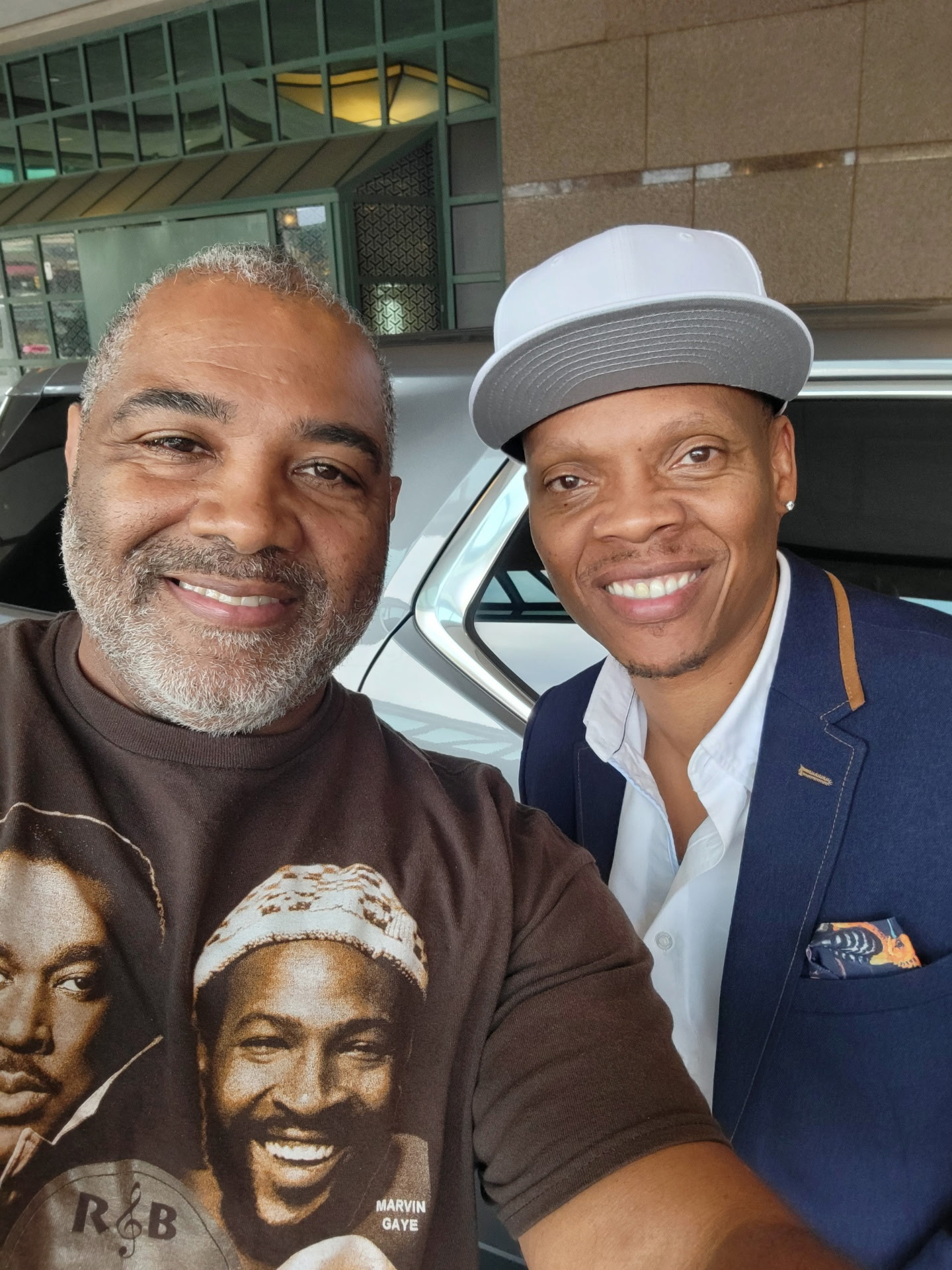 Two men smiling, one wearing a graphic T-shirt and the other in a suit with a hat, standing outside a building.