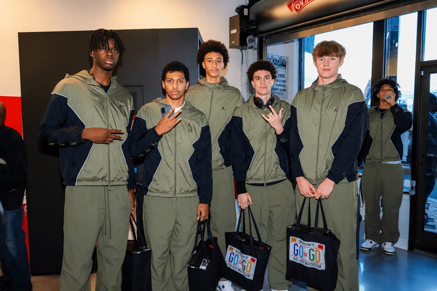 Five young men stand indoors, wearing matching green and black jackets, holding bags, with a glass door behind them.
