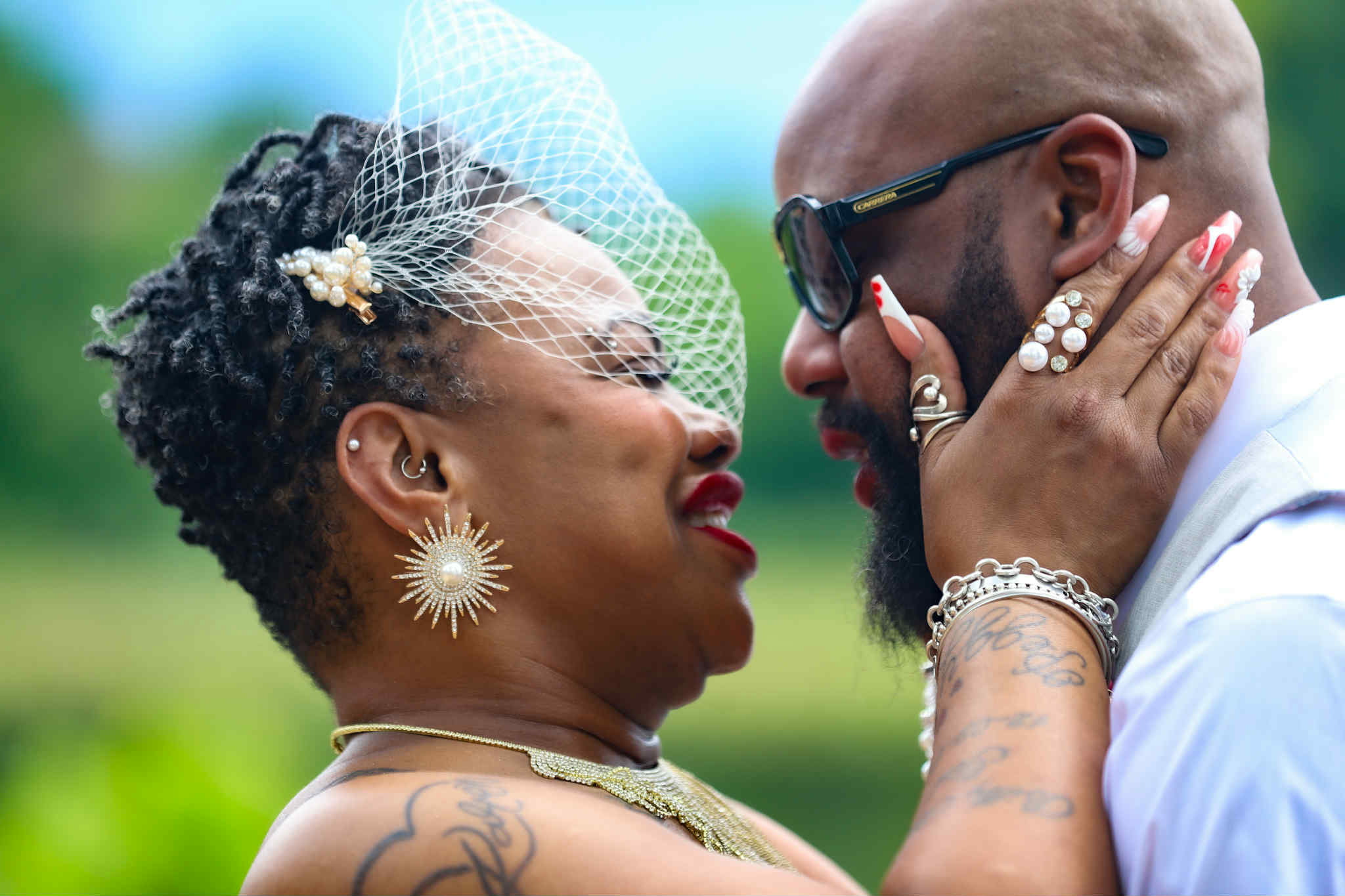A woman and man face each other closely outdoors, holding each other's faces, smiling, with a blurred green background.