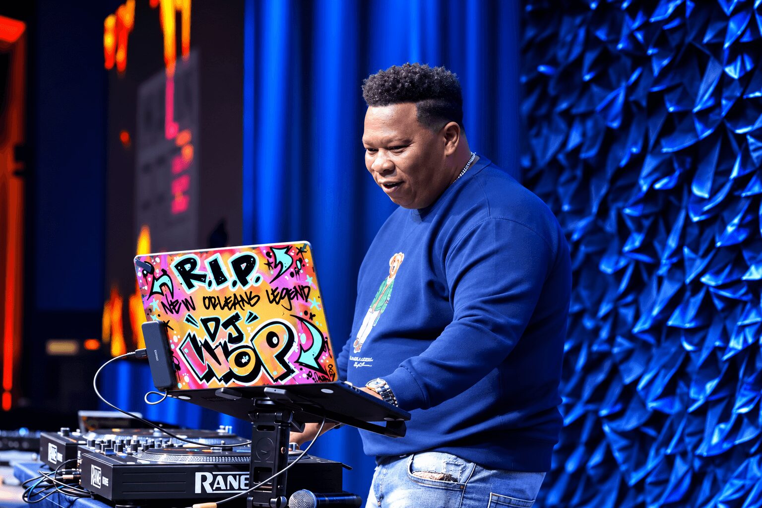 Person in blue sweatshirt using a laptop with colorful stickers, standing in front of a blue textured backdrop.