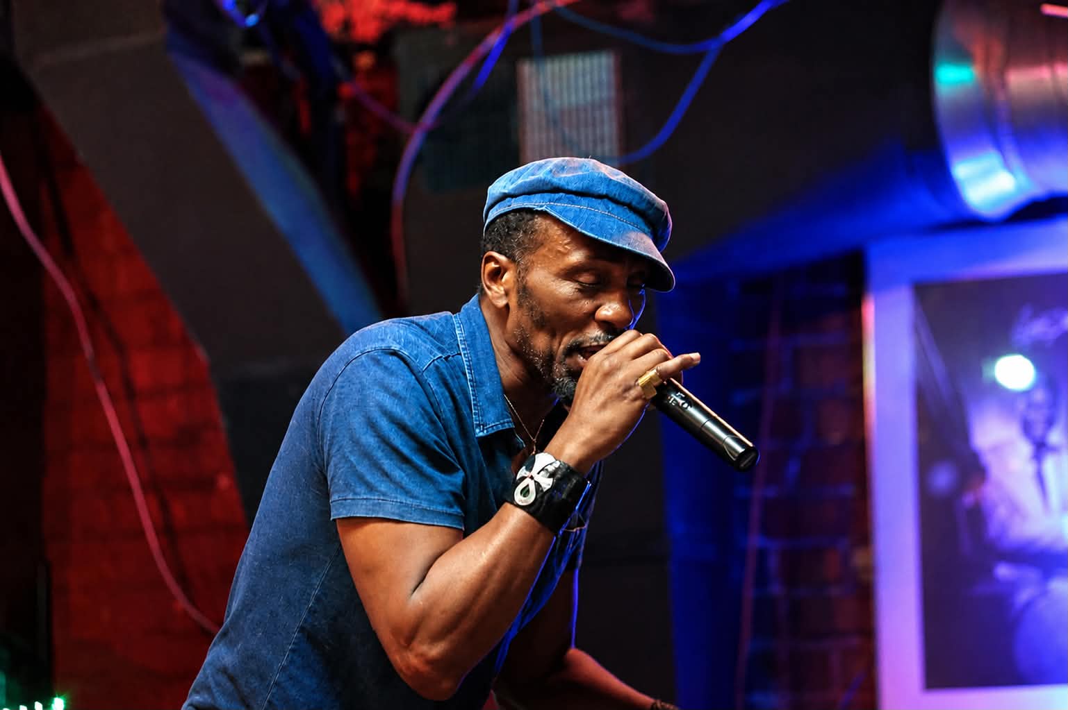 Man singing into a microphone on stage, wearing a blue cap and shirt, with colorful stage lighting.