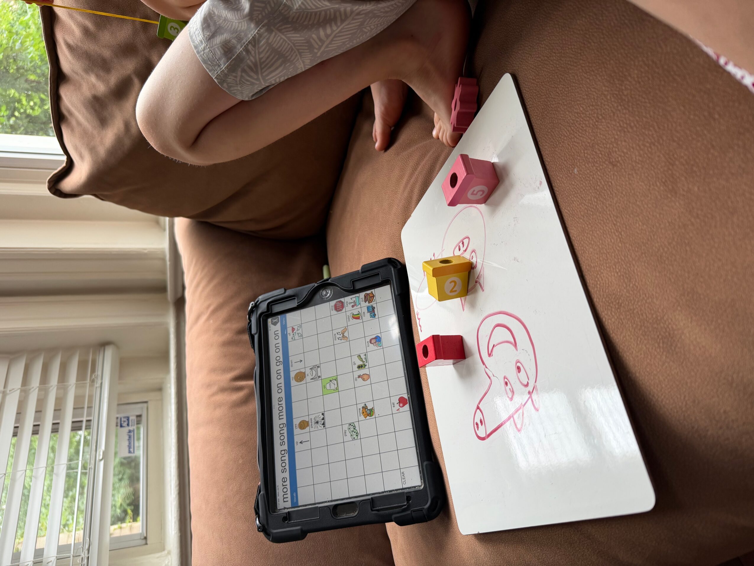 Person drawing a face on a whiteboard with markers, tablet displaying a grid, and a window with blinds in background.