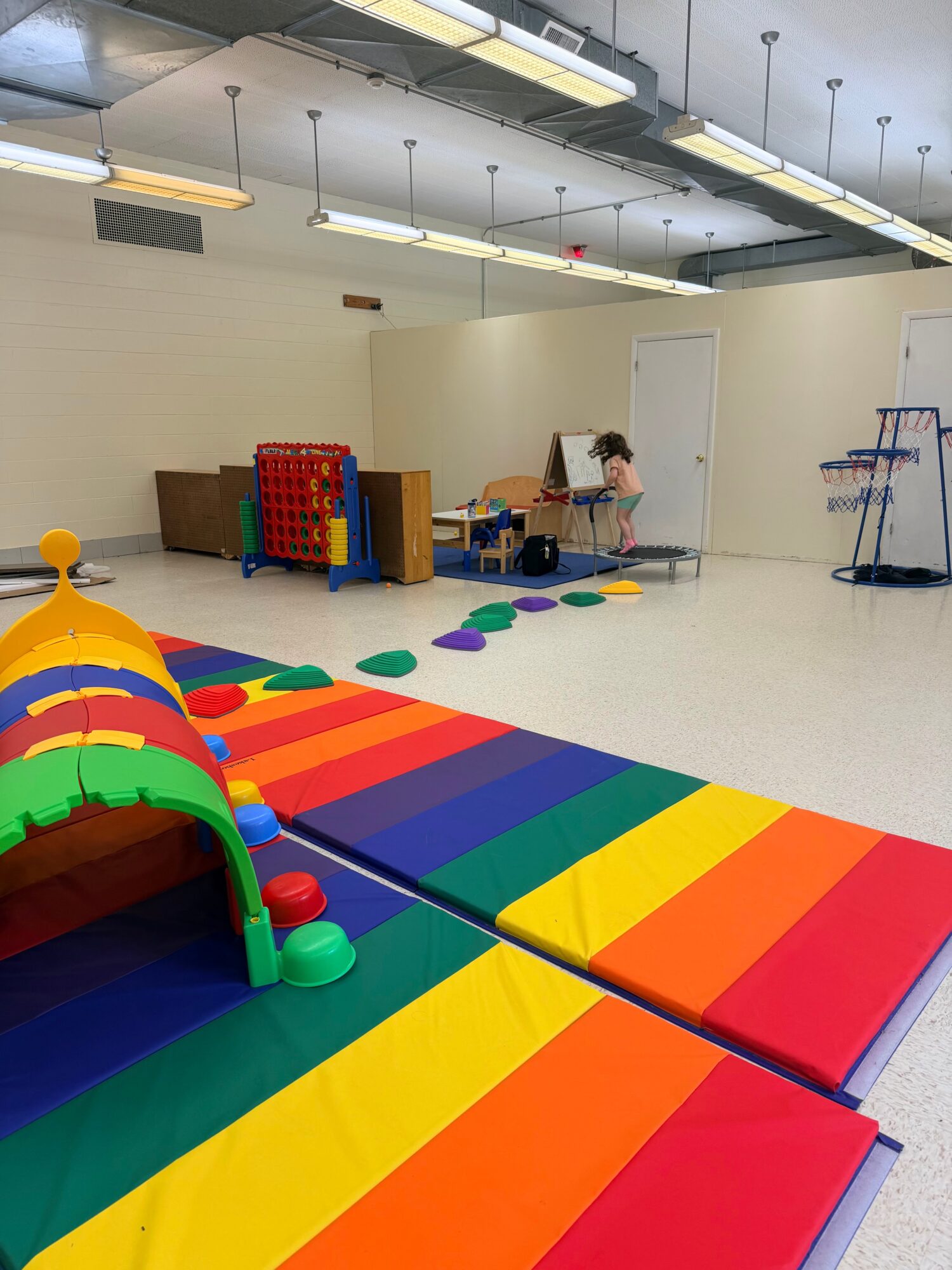 Indoor play area with colorful mats, a small table, and a person sitting at the table, with a white wall and ceiling lights.
