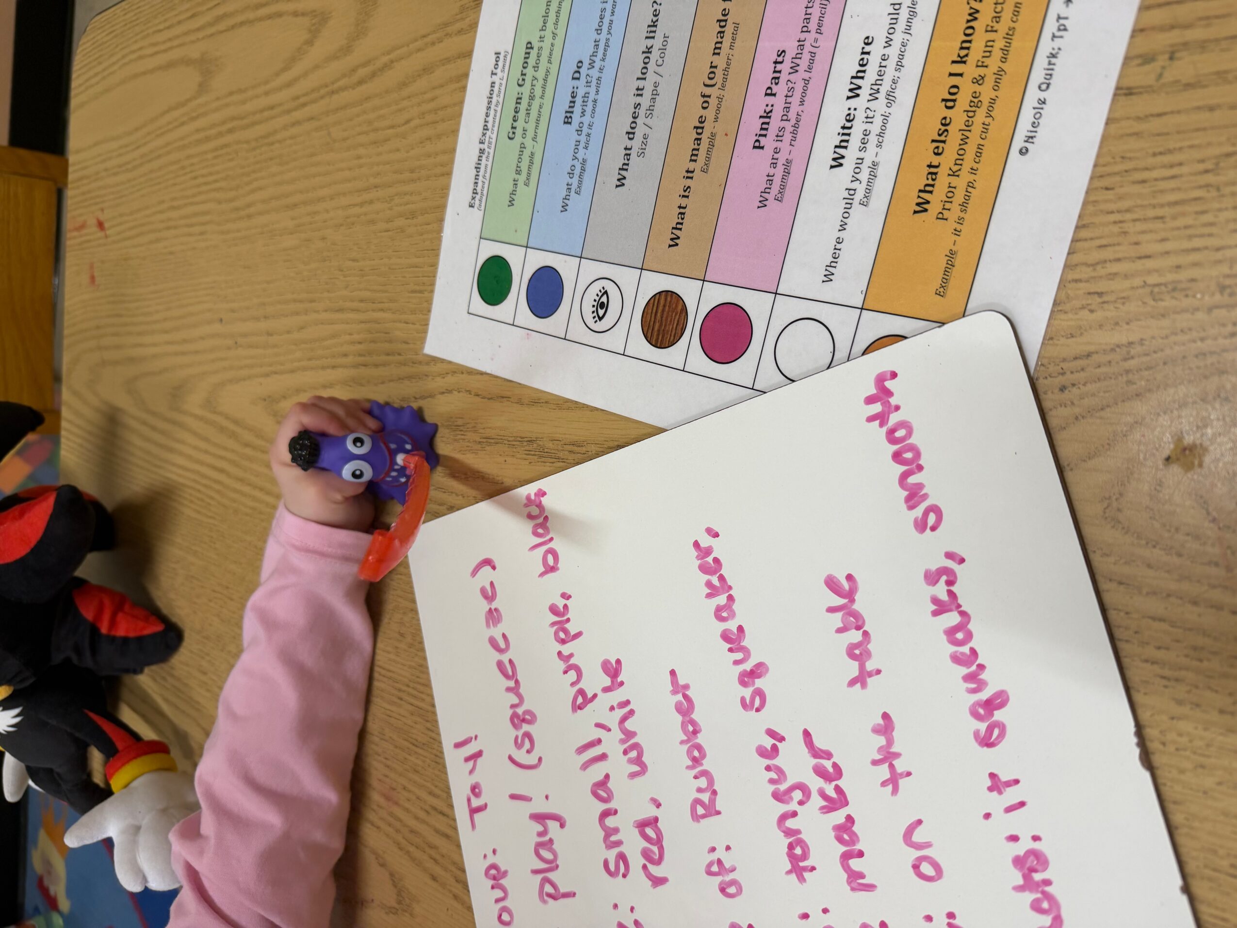 A child's hand holding a purple crayon, writing on a whiteboard with pink marker. A colorful chart is on the table.