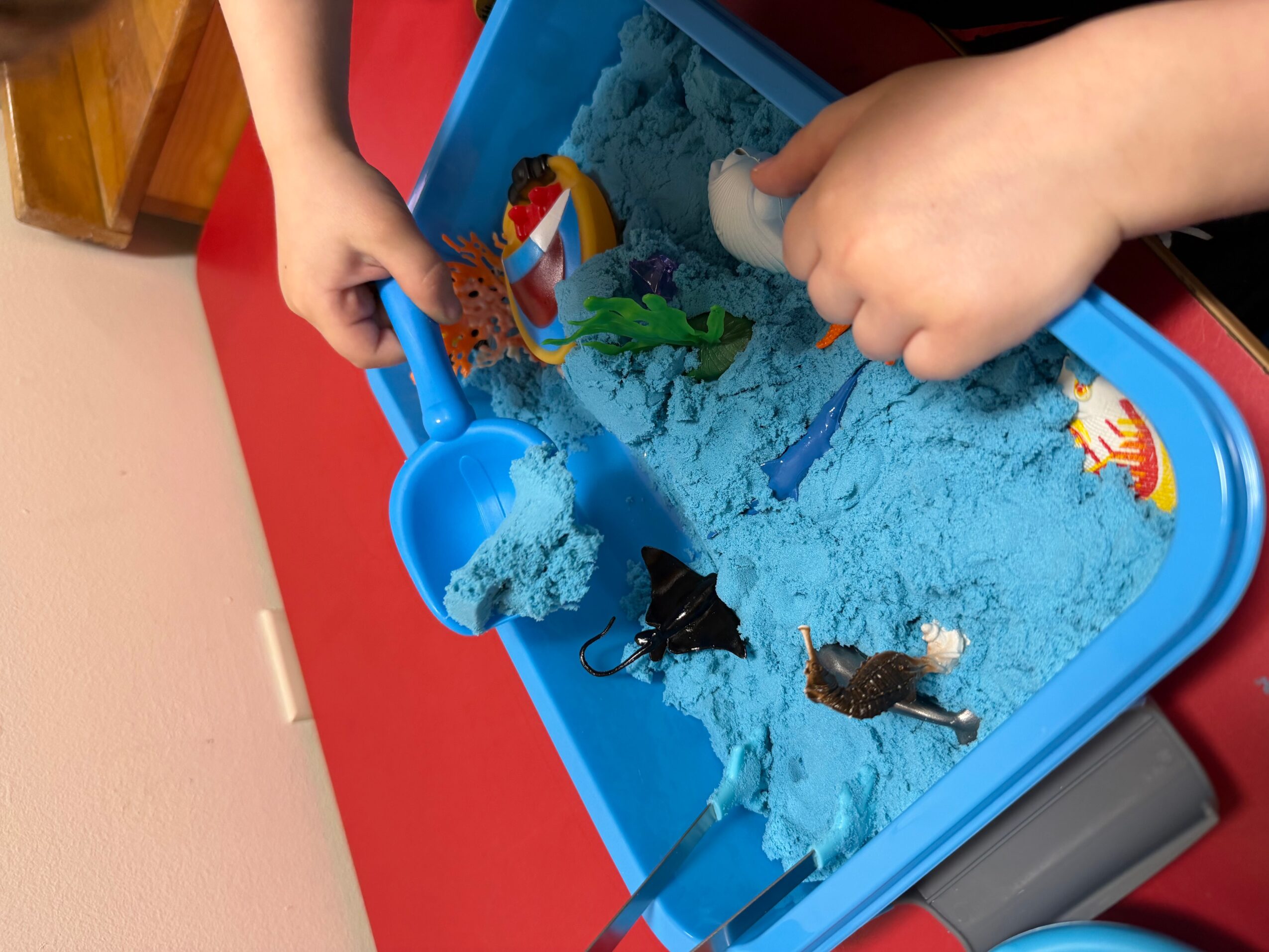 Two children play with a blue sand toy tray filled with blue sand, using their hands and a small shovel, with toy sea creatures inside.