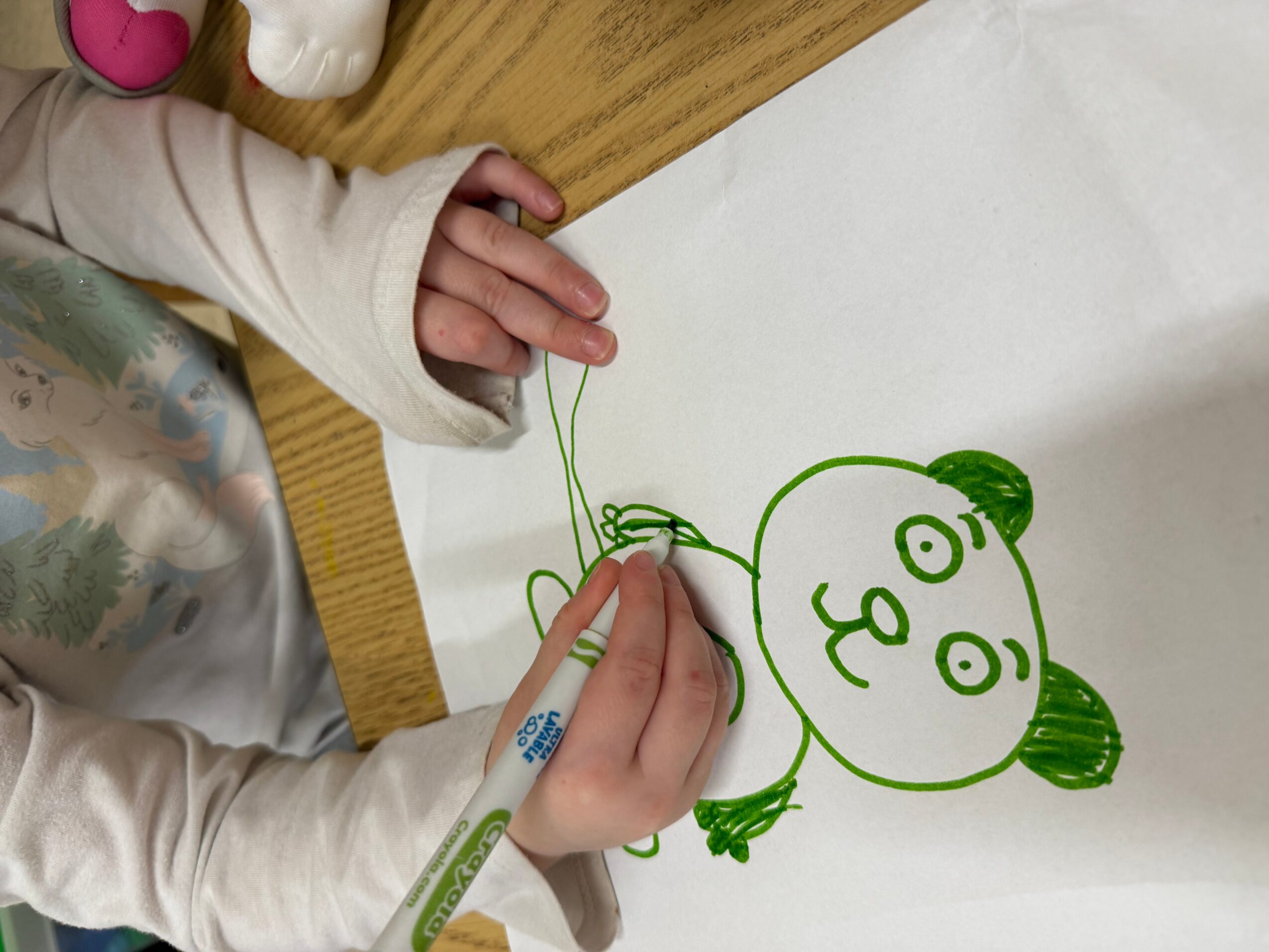 Child drawing a cartoon bear face with a green marker on white paper, with a wooden table nearby.