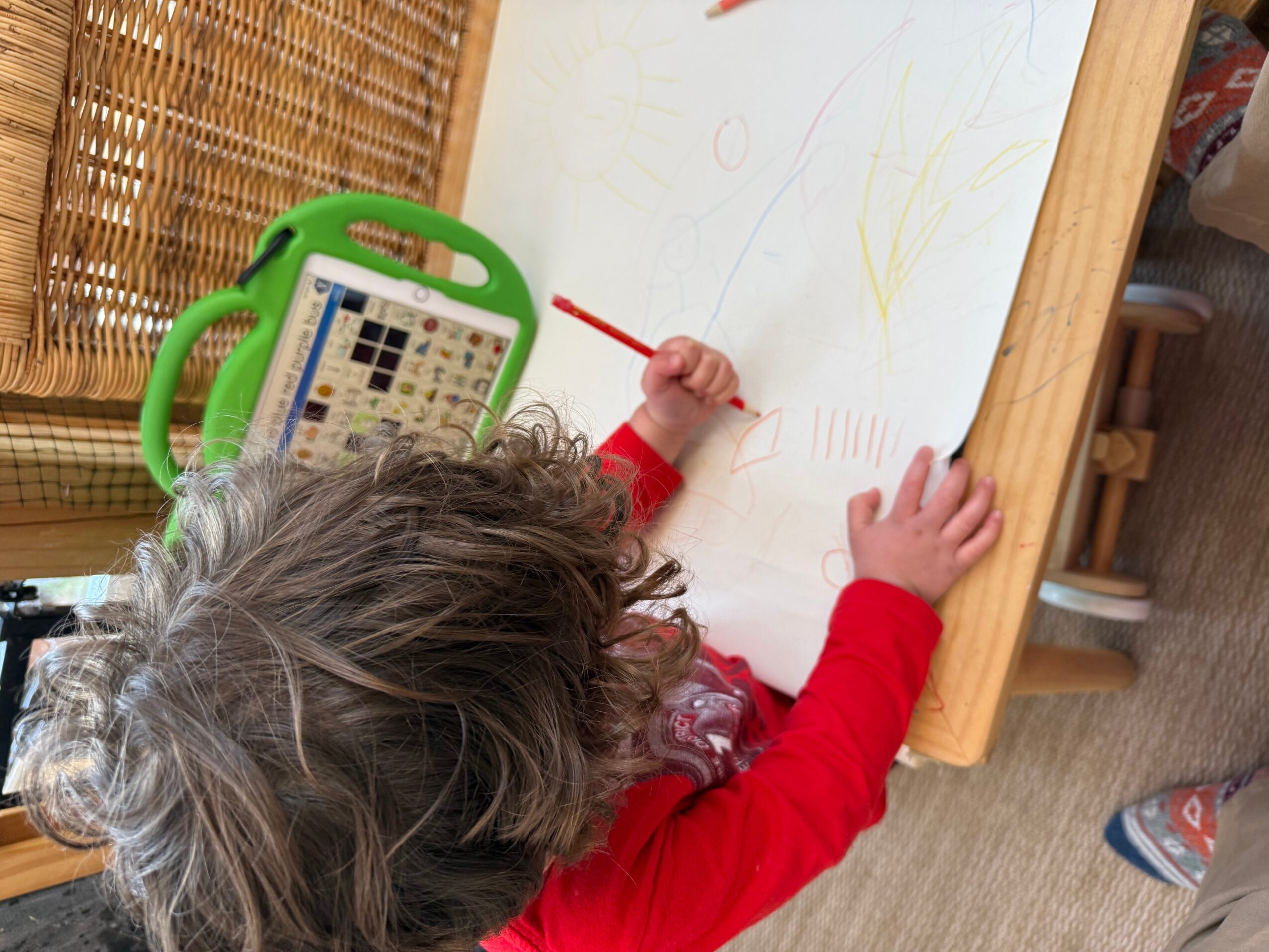 Child drawing on a whiteboard with a red marker, wearing a red long-sleeve shirt, with a green electronic device nearby.