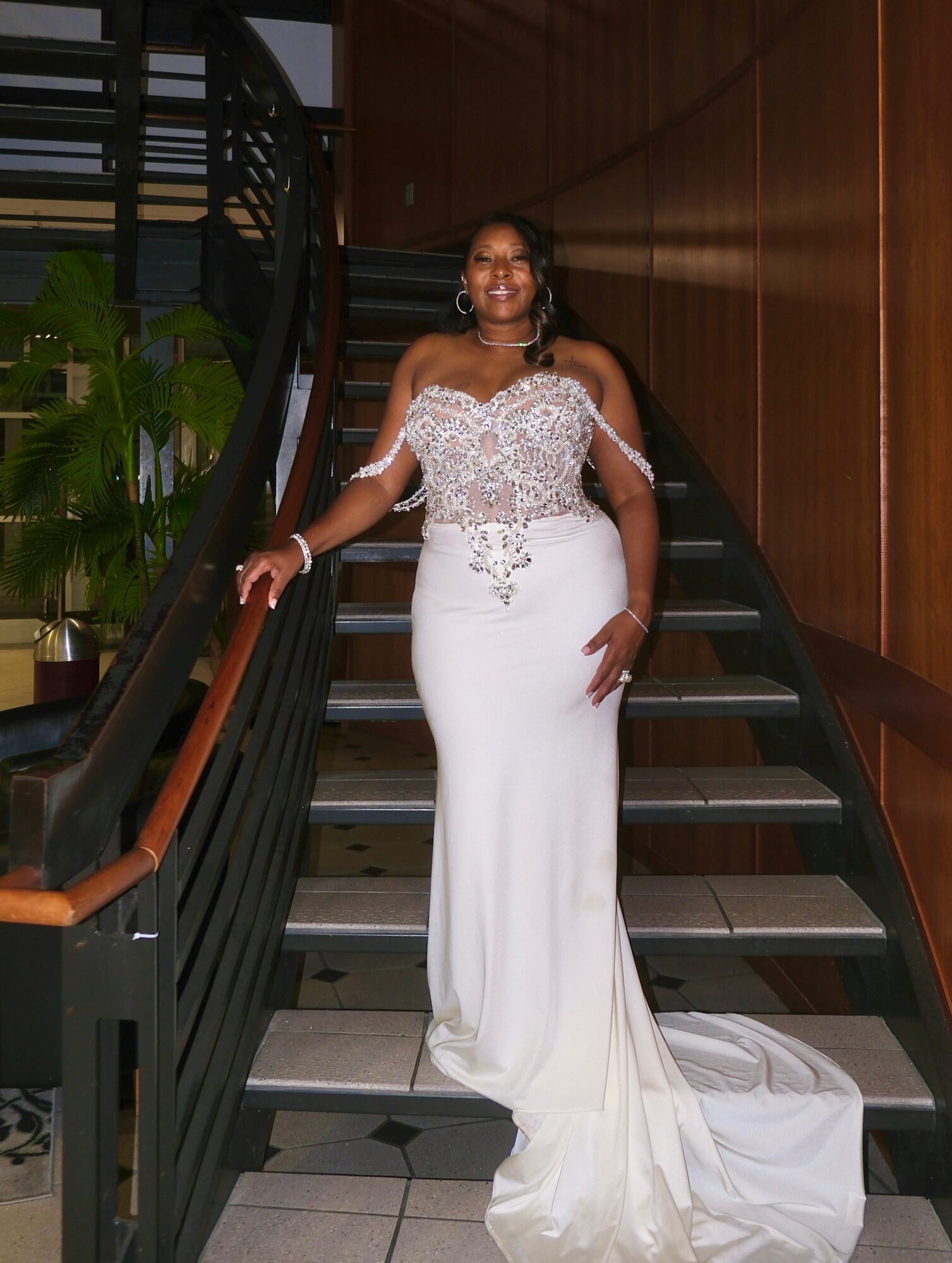 Woman in a white wedding dress standing on a staircase indoors, smiling, with wooden walls and green plants nearby.
