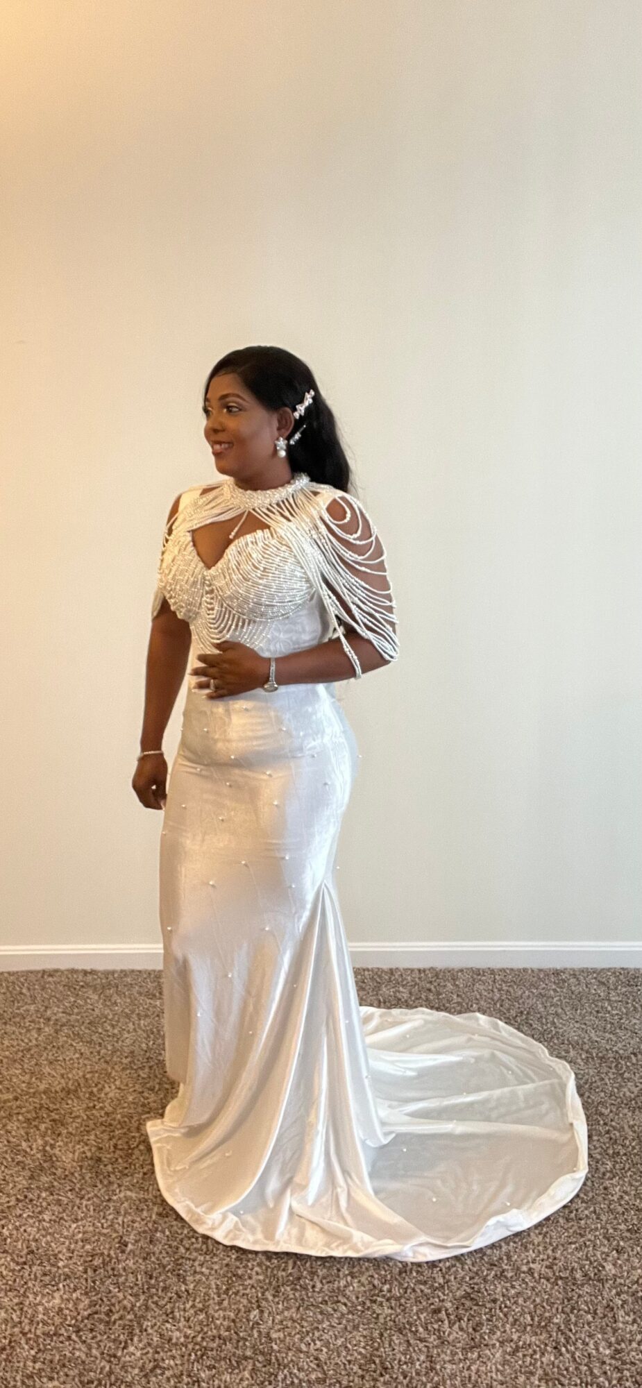 Woman in a white wedding dress standing on a carpeted floor against a plain wall, with her hands clasped in front.