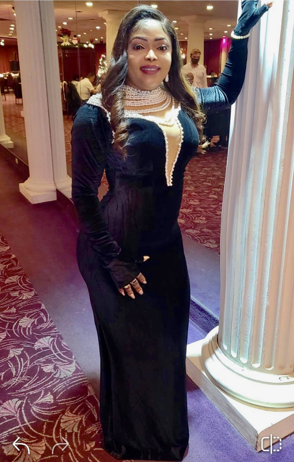 Woman in elegant black dress with pearl necklace, standing next to a white column in a decorated indoor setting.