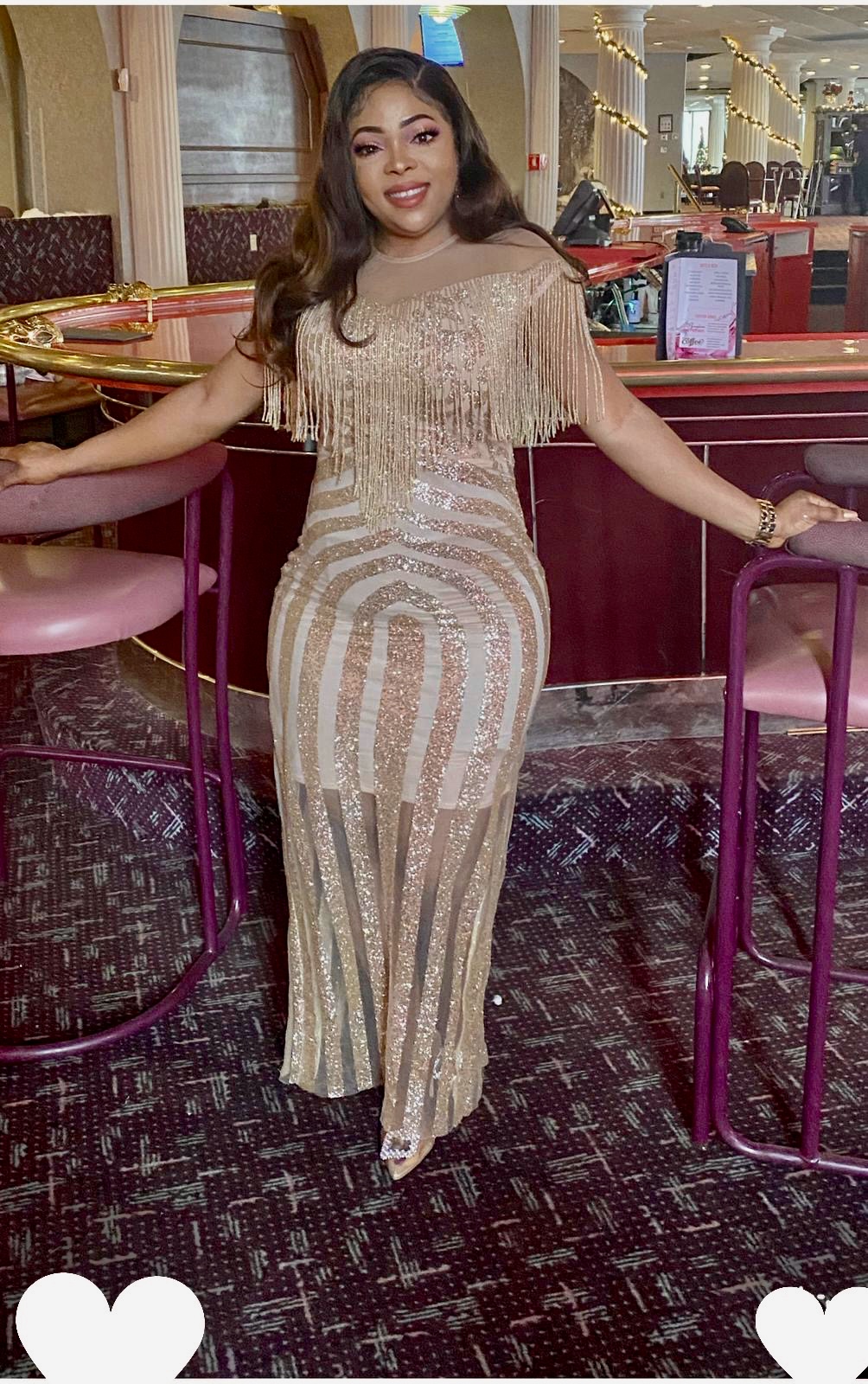 Woman in a beige, sequined dress standing with arms outstretched in a restaurant or lounge setting.